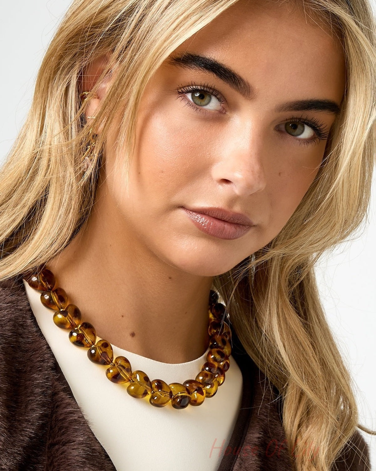 Close-up front view of woman wearing amber heart bead necklace paired with cream top and brown vest