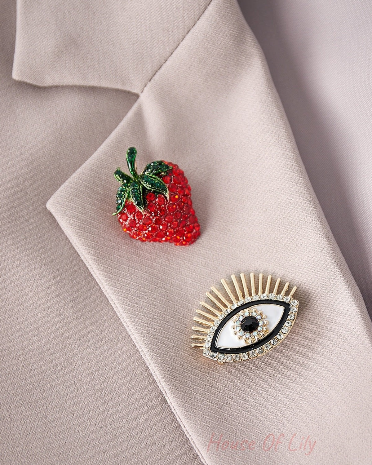 Close-up of beige blazer lapel decorated with a red rhinestone strawberry brooch and gold-tone eye brooch with rhinestones