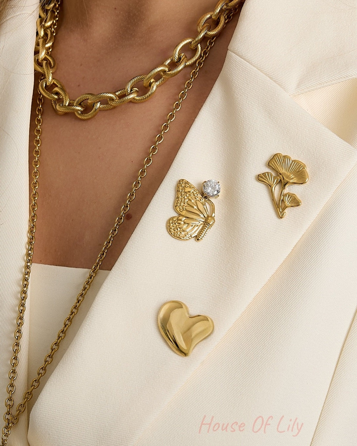Woman wearing cream blazer adorned with three gold brooches: butterfly with rhinestone, floral design, and heart, paired with gold chain necklaces.