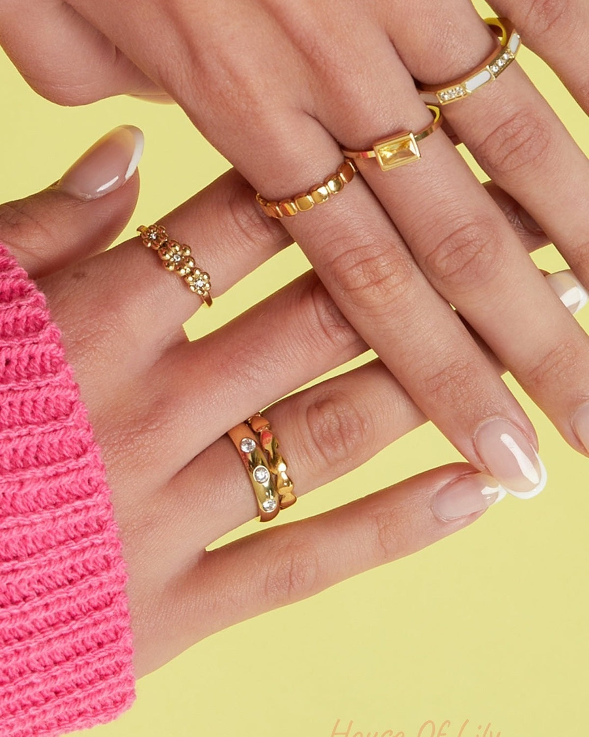 Close-up image of a woman's hand wearing multiple gold rings including a heart link ring on yellow background with pink sweater sleeve
