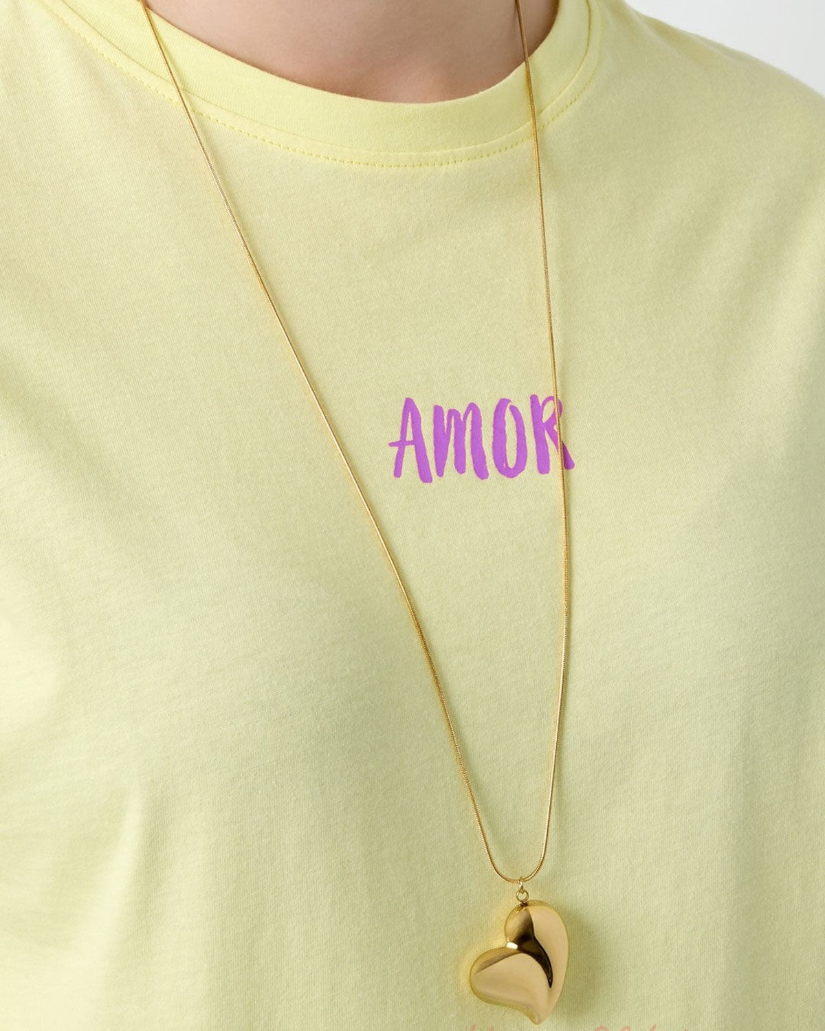 Gold heart pendant necklace worn over a pastel yellow t-shirt with the word 'AMOR' printed in purple