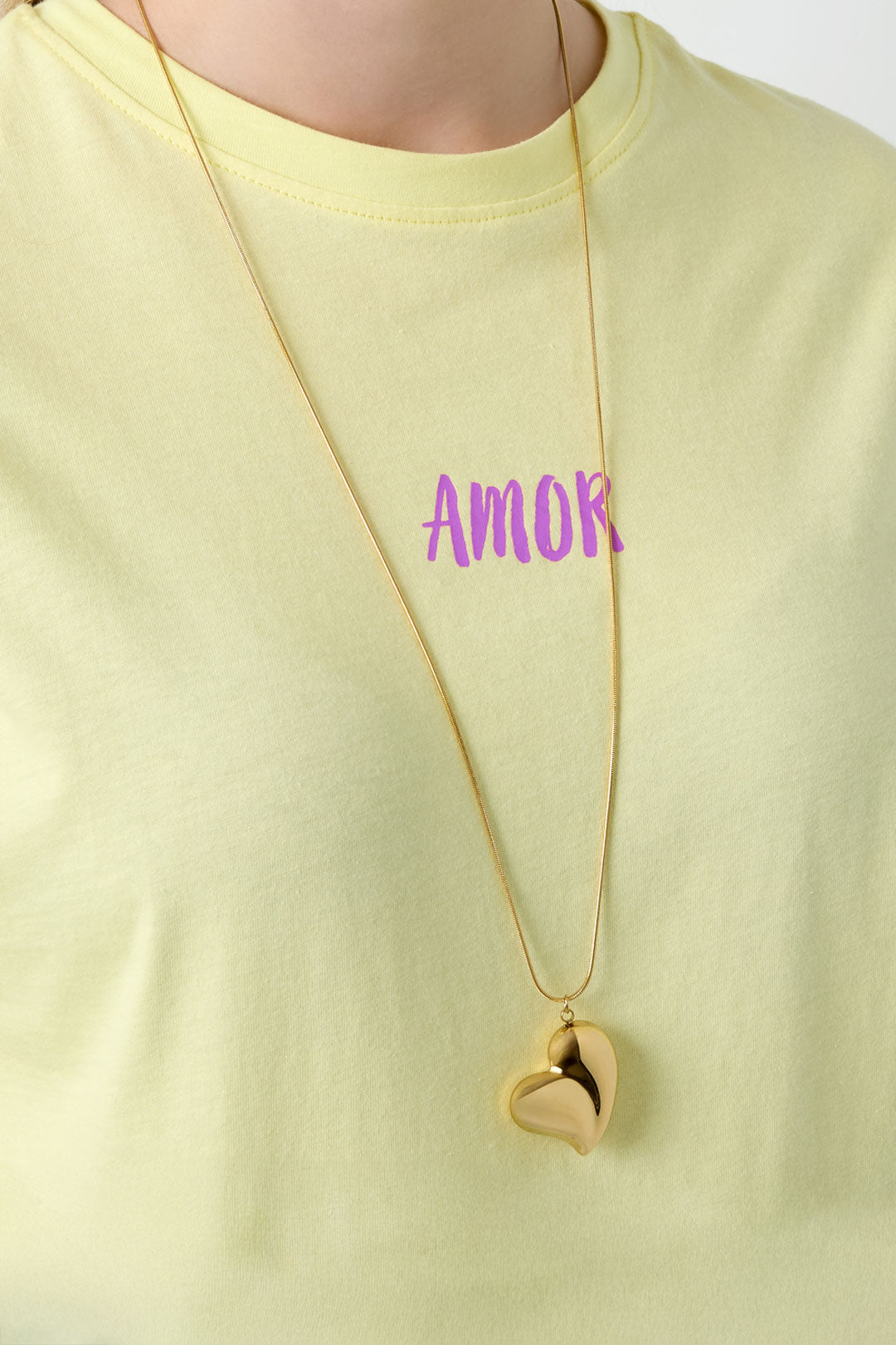 Gold heart pendant necklace worn over a pastel yellow t-shirt with the word 'AMOR' printed in purple