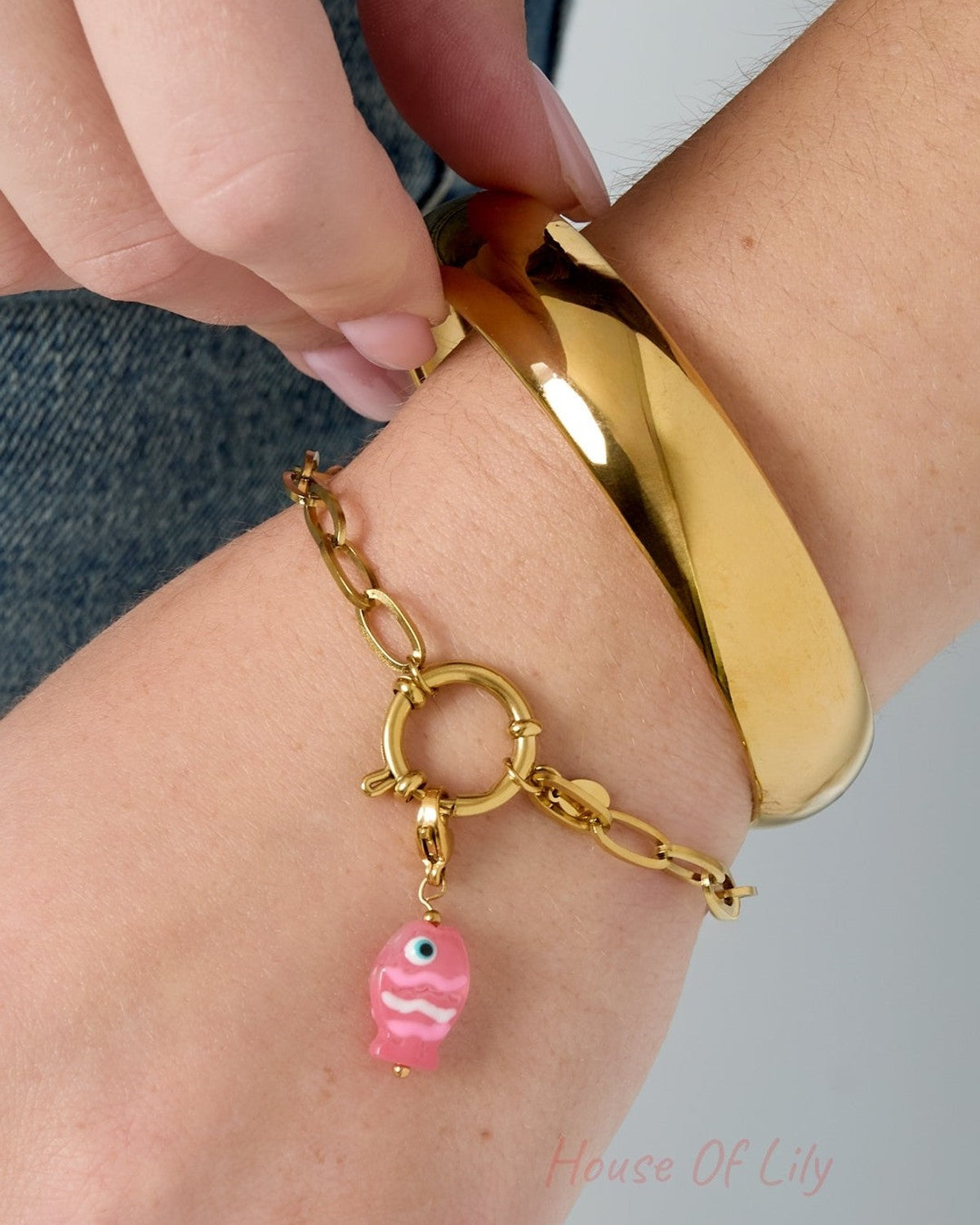 Wrist wearing a gold chain bracelet with a pink fish charm alongside a gold bangle