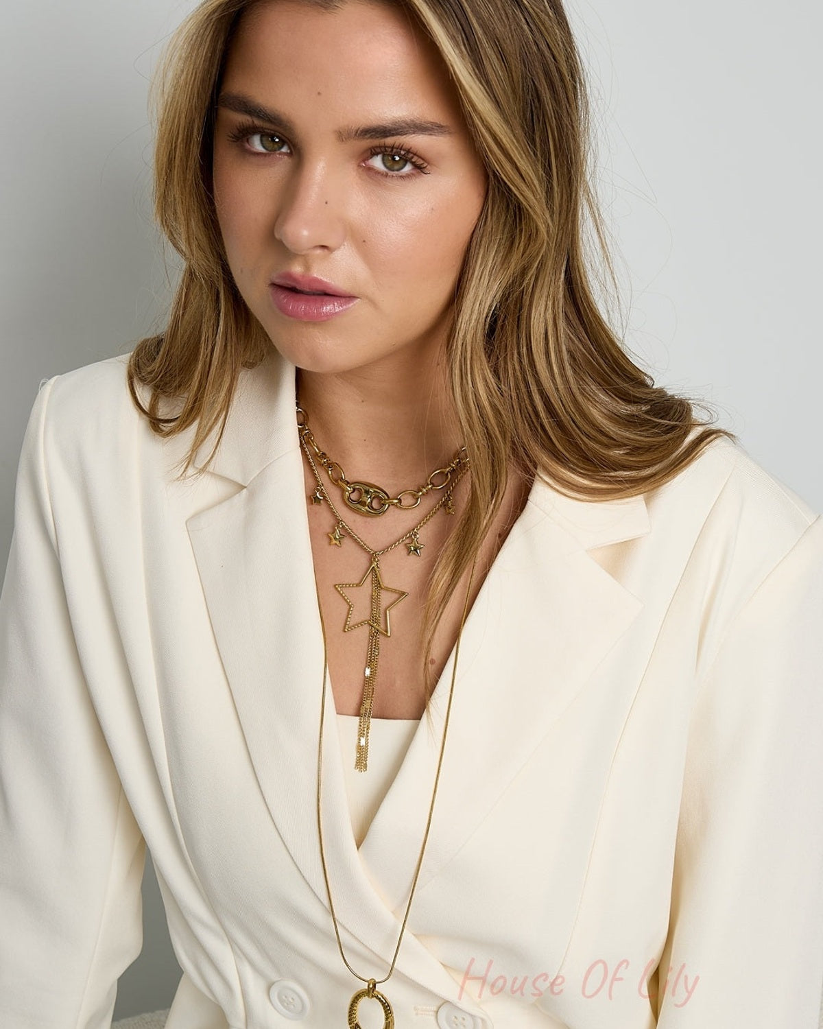 Model wearing gold star charm necklace layered with chunky chain and long gold pendant necklace with cream blazer