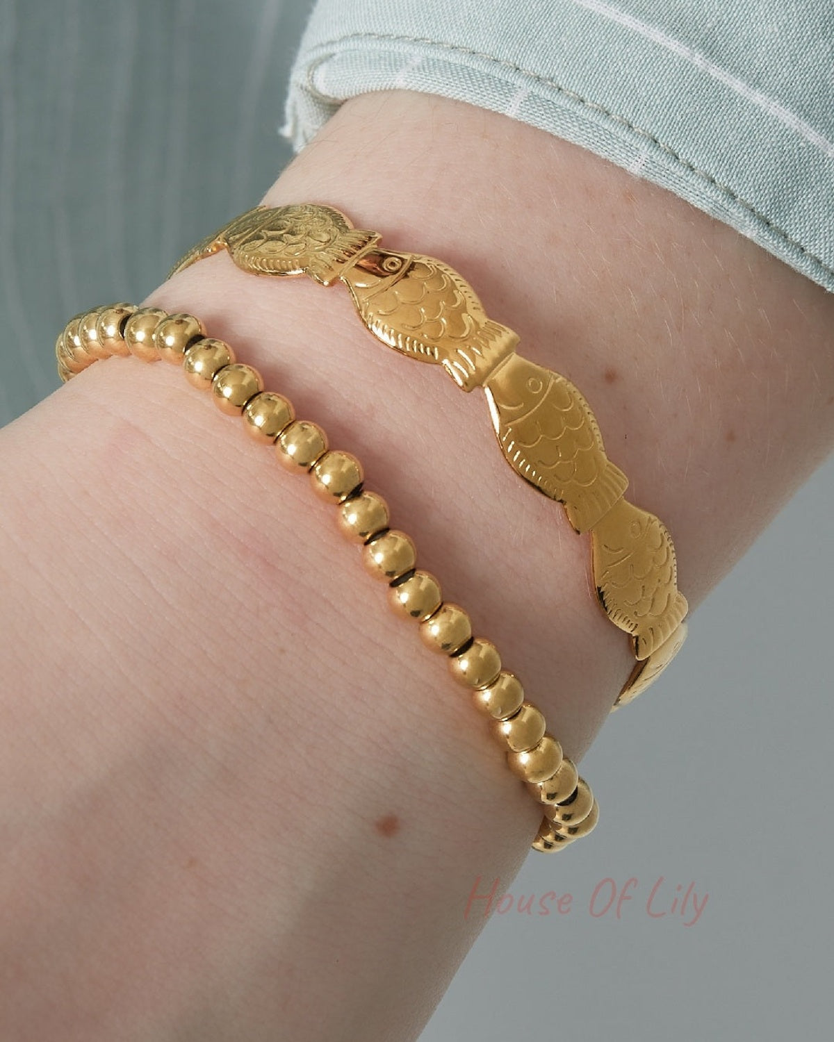Close-up of wrist wearing gold beaded stretch bracelet paired with a gold fish-design bracelet and light blue sleeve