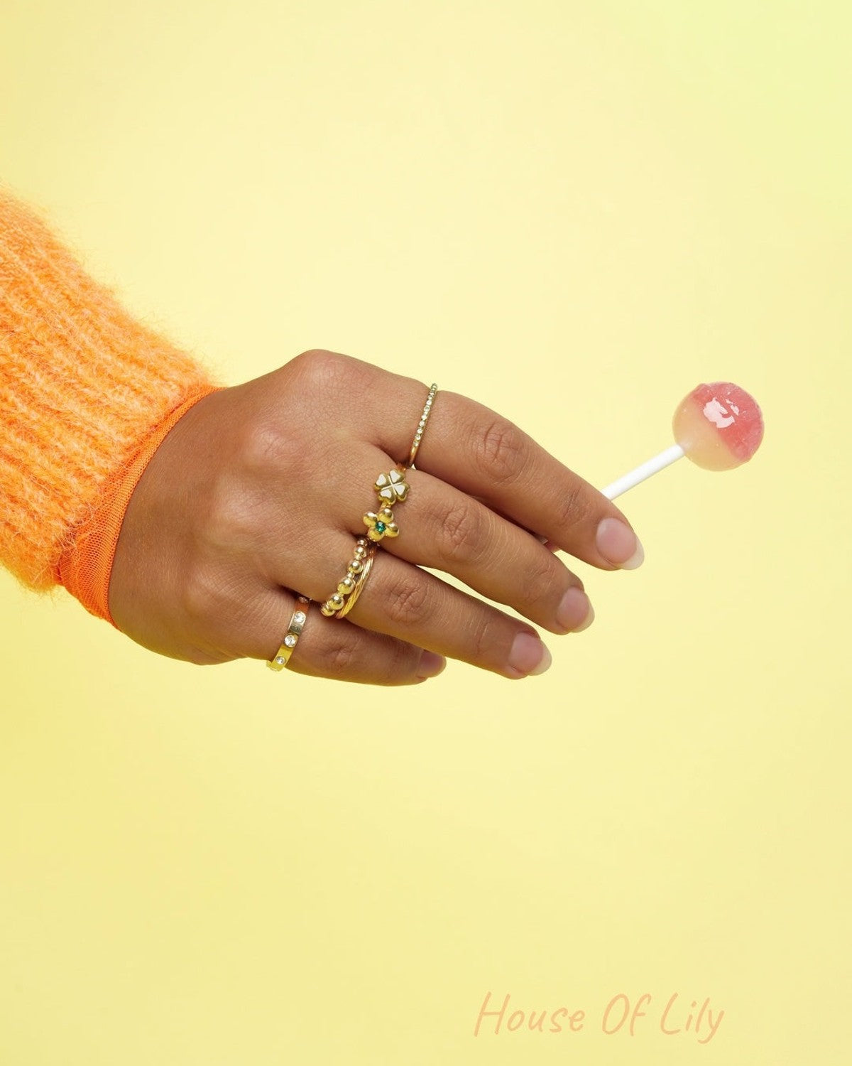 Woman's hand with multiple gold rings holding a pink and white lollipop against yellow background