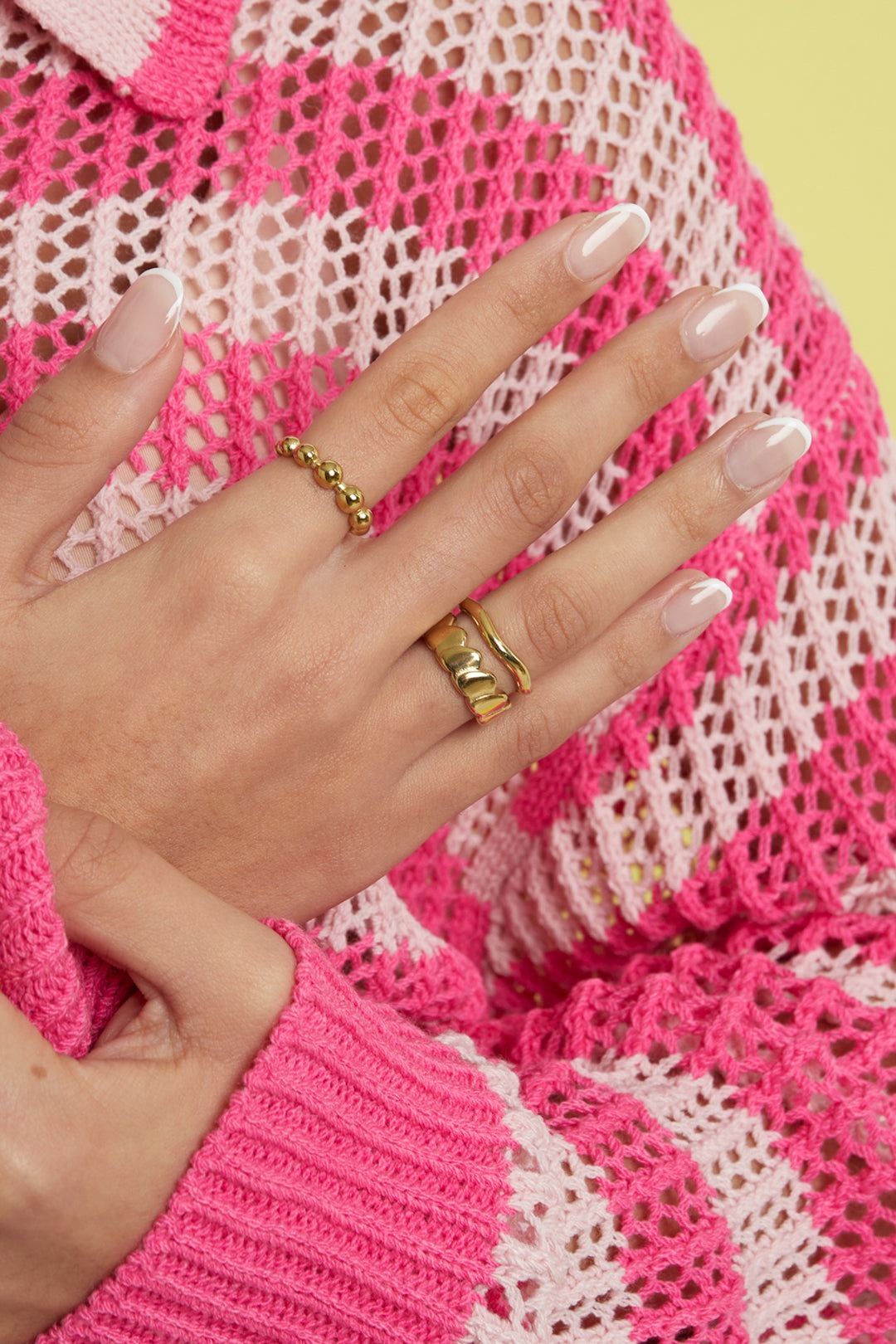 Female hand wearing a gold heart chain ring with two other gold rings, showcasing polished nails and pink knitwear background