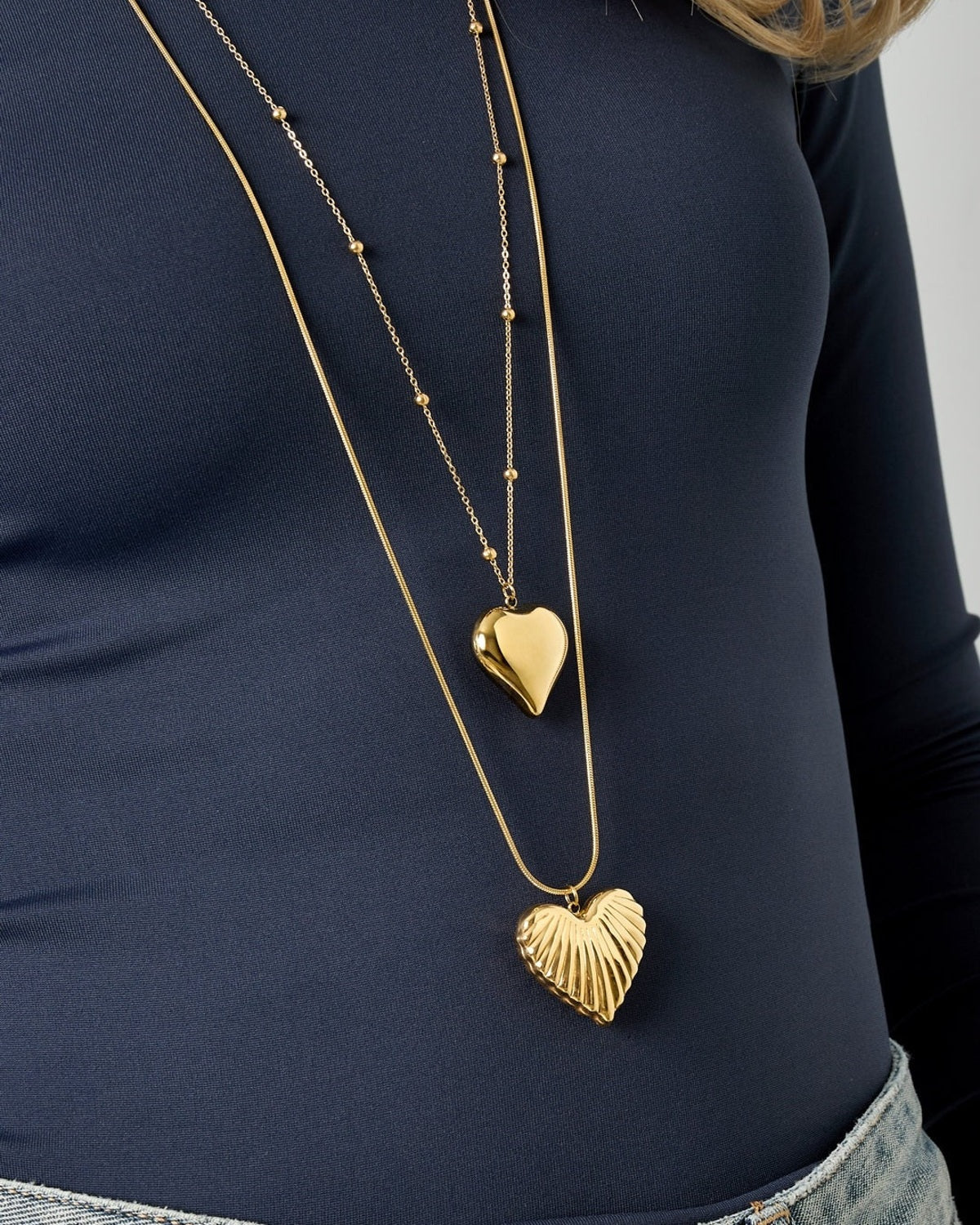 Close-up shot of the layered gold heart pendant necklaces showcasing the textured heart on light-wash denim and navy blue top