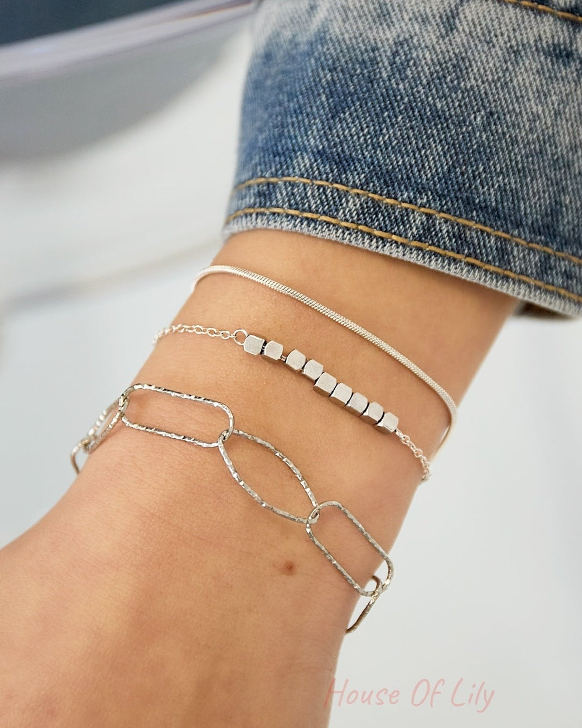Close-up of silver-tone dainty cube chain bracelet layered with two other bracelets on wrist wearing denim sleeve