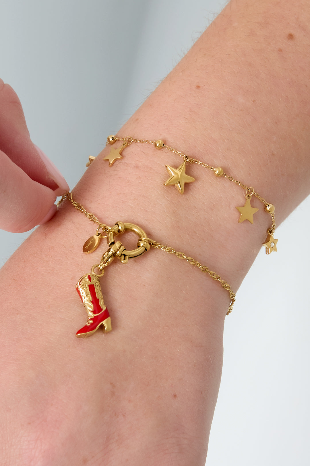 Close-up of wrist wearing two gold bracelets including chain toggle bracelet with red cowboy boot charm and star charm bracelet