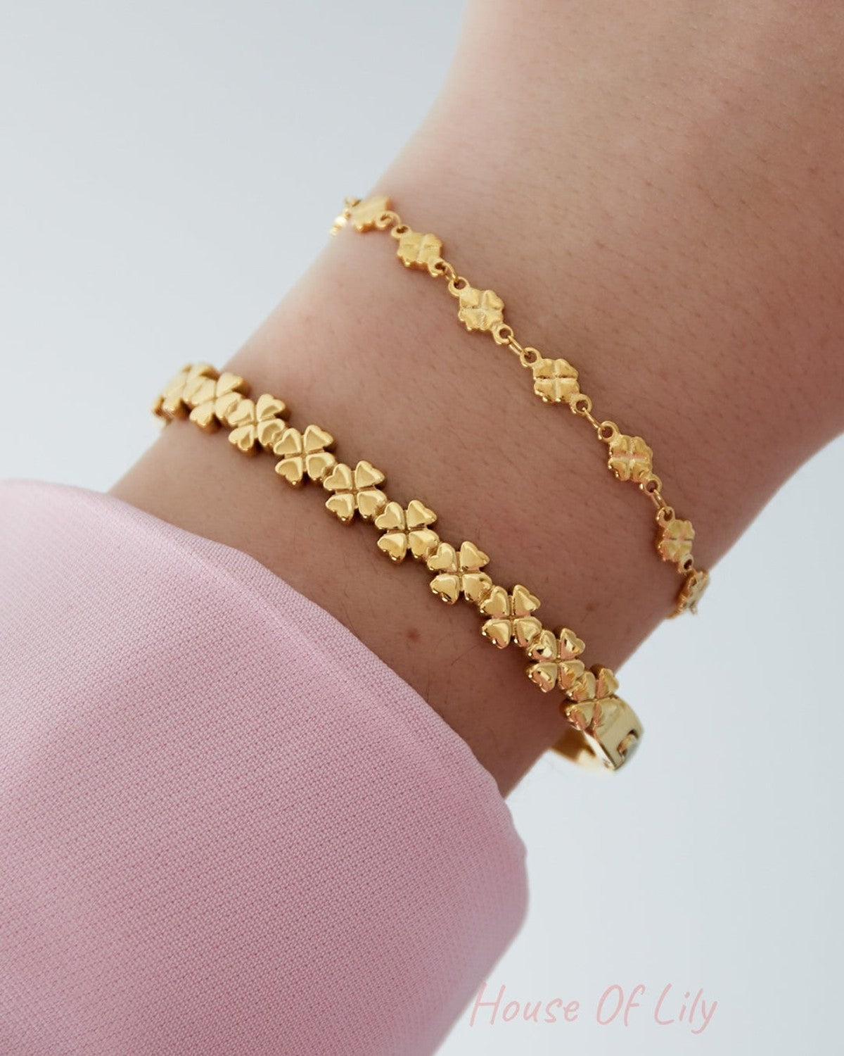 Close-up of two gold four-leaf clover bracelets worn stacked on a wrist with pink sleeve