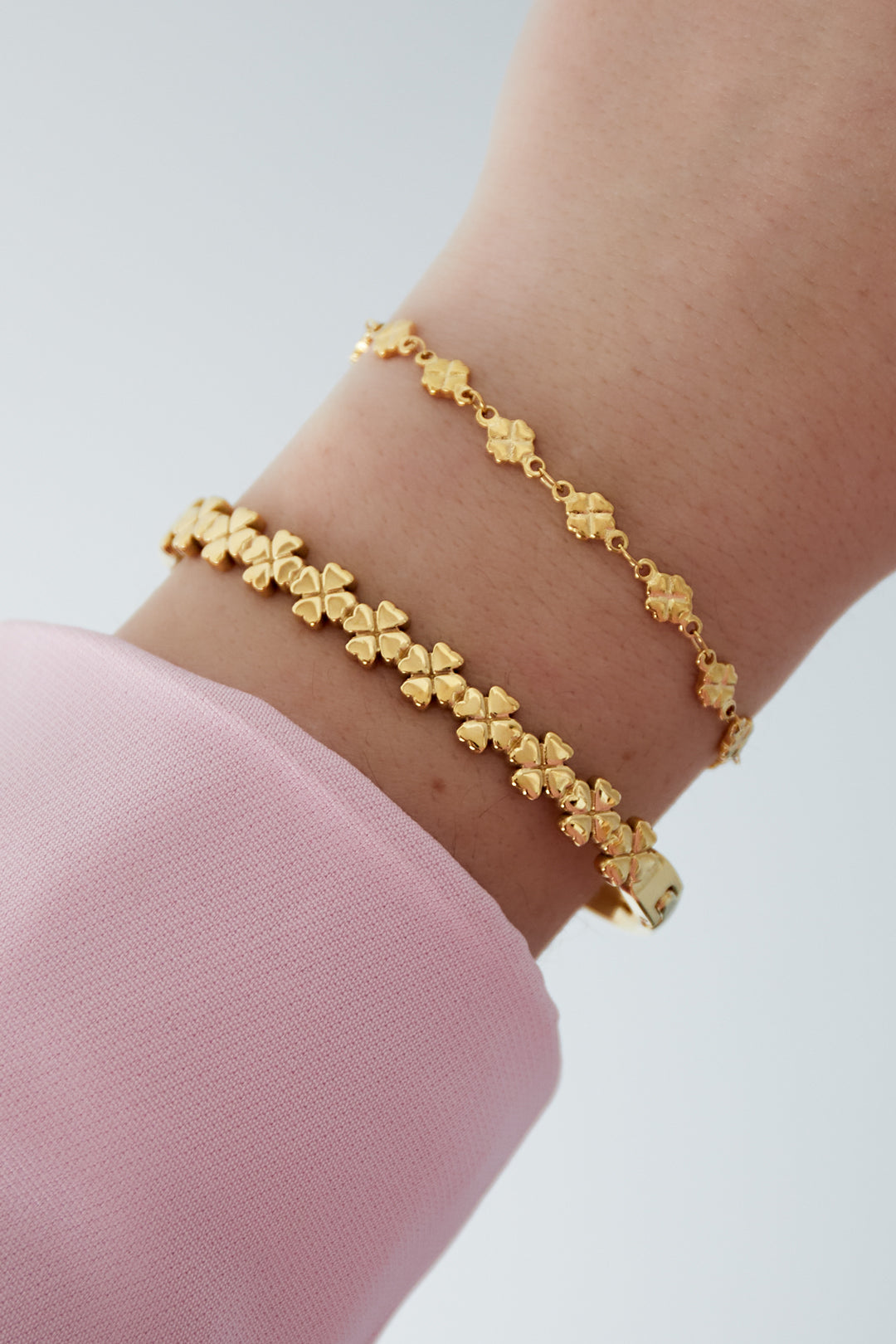 Close-up of two gold four-leaf clover bracelets worn stacked on a wrist with pink sleeve