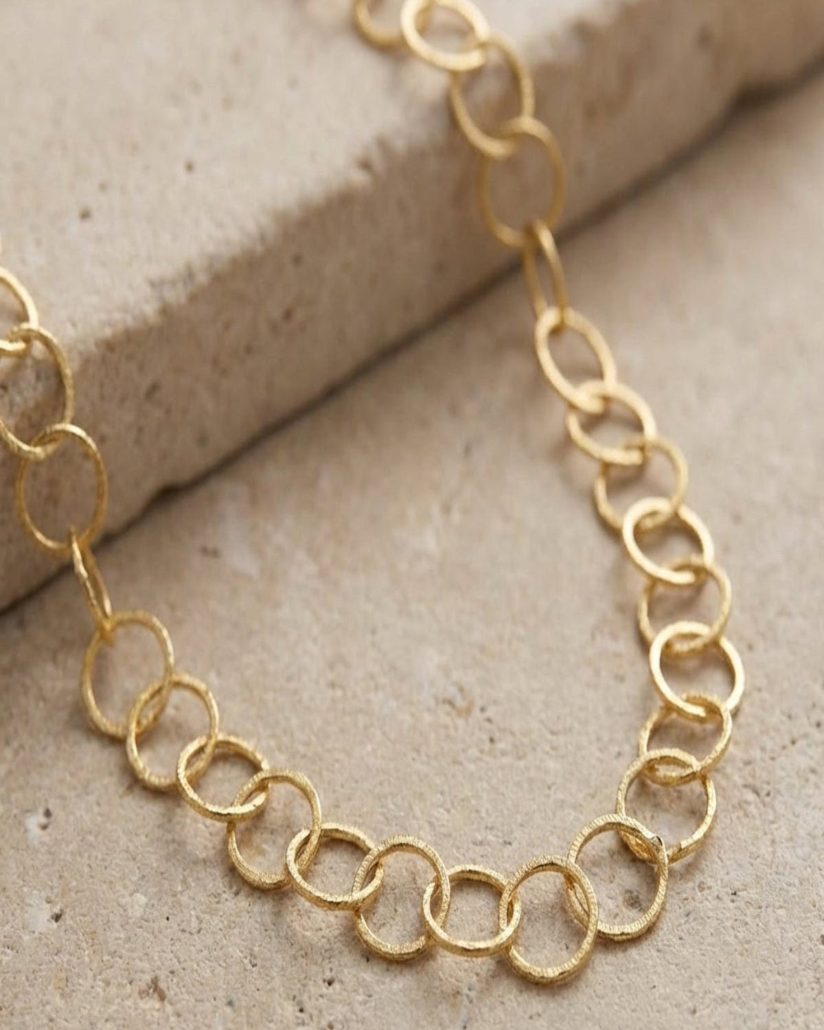 Close-up of a delicate gold-plated stainless steel loop chain necklace with fine links, showcased on a neutral textured surface with soft lighting.