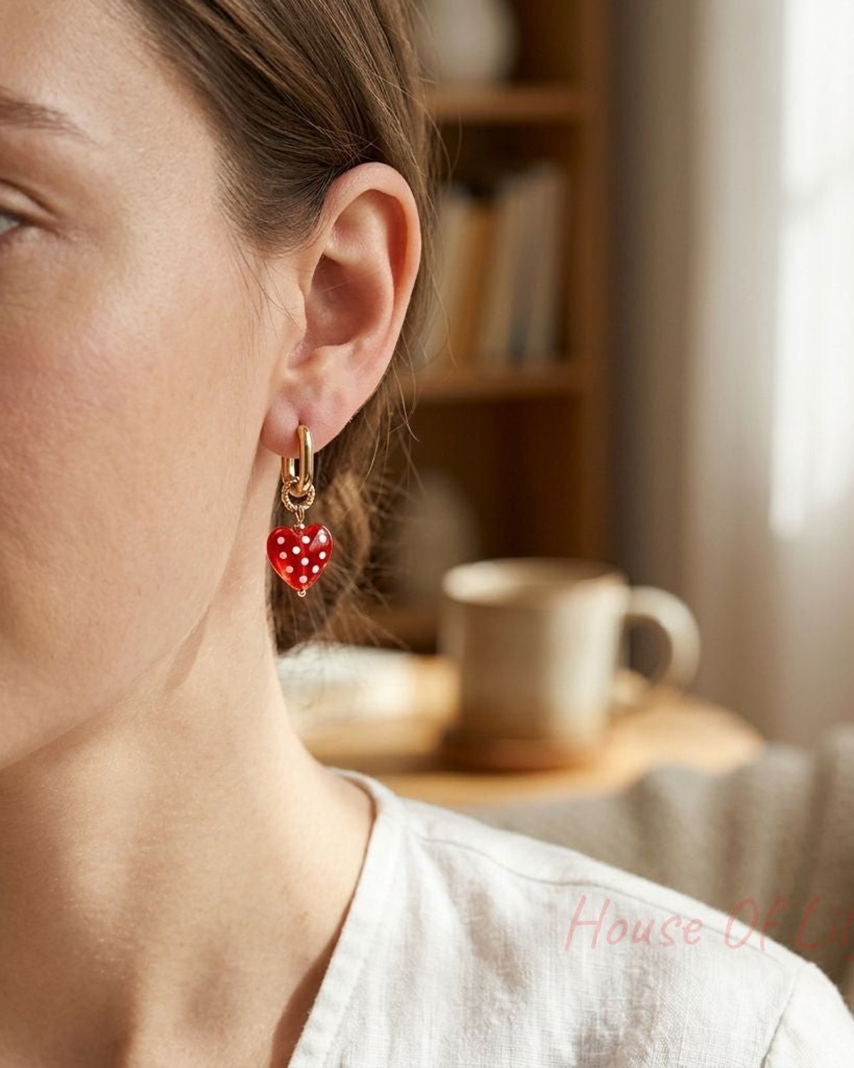Woman wearing dotted heart drop earrings in cozy indoor setting, showcasing small, feminine jewelry with natural sunlight.