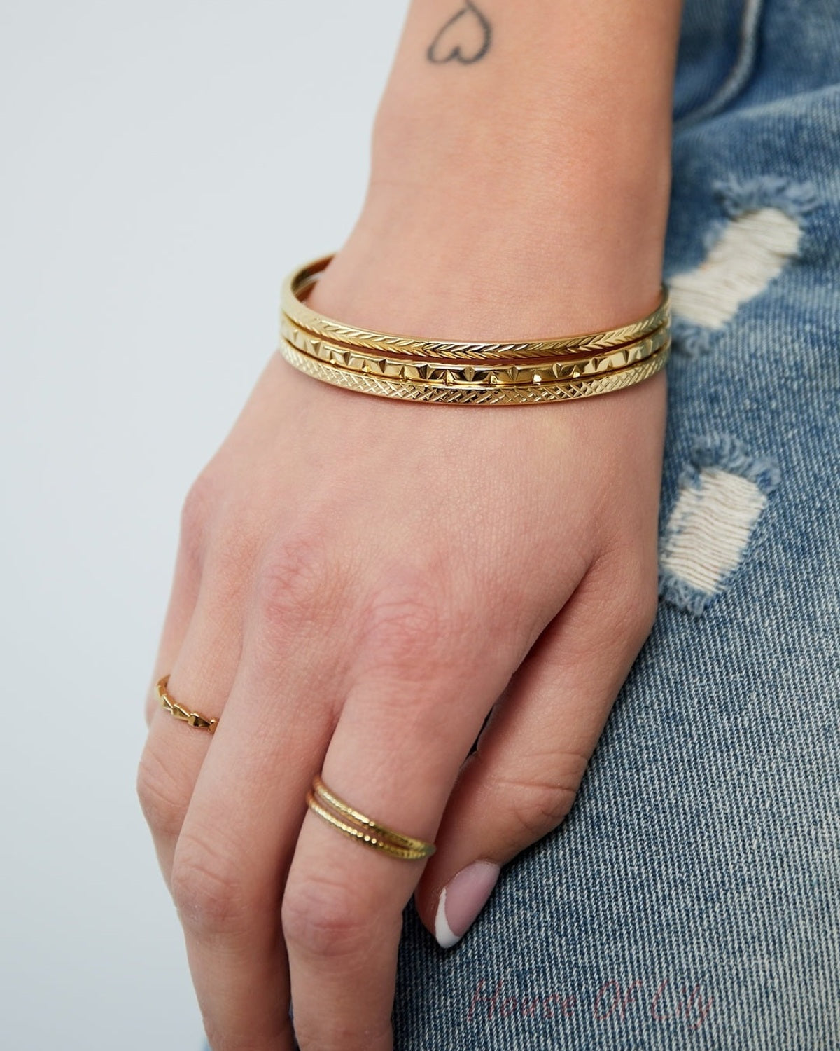 Woman's hand resting on denim-clad leg, wearing a set of three gold-tone slim bangles and matching rings