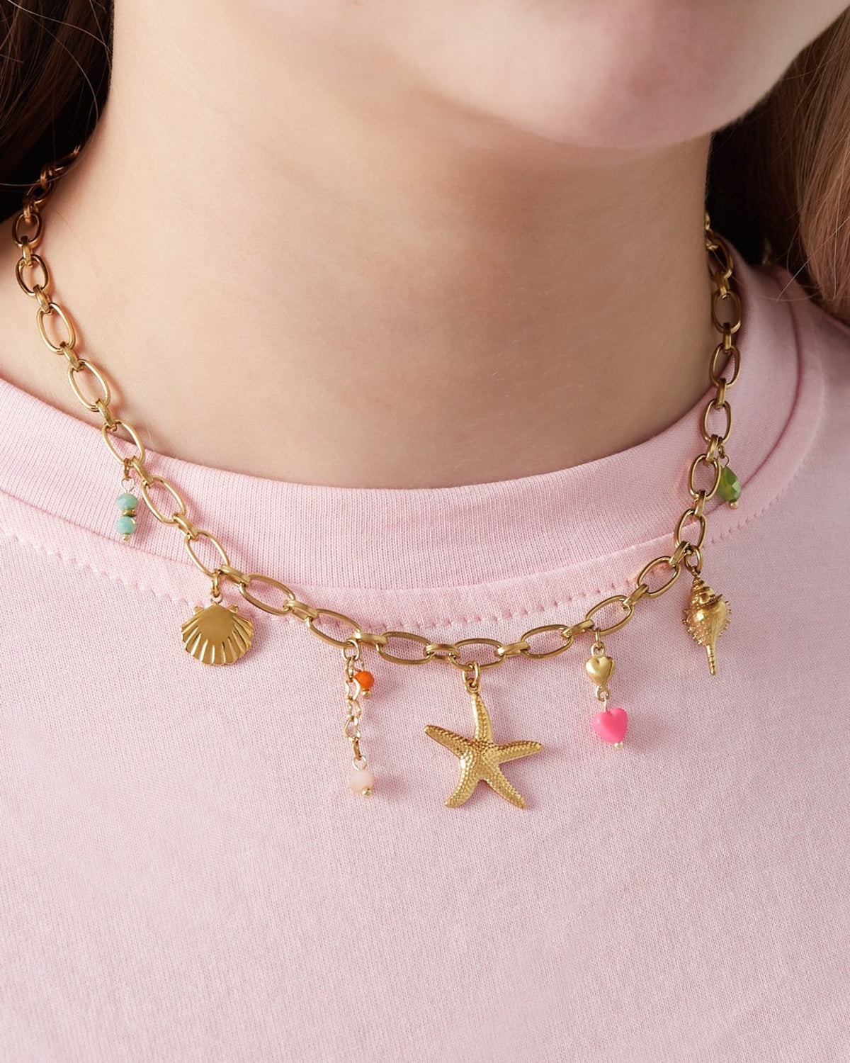 Close-up of gold-tone charm necklace worn by woman with pastel pink top, showing beach-themed charms