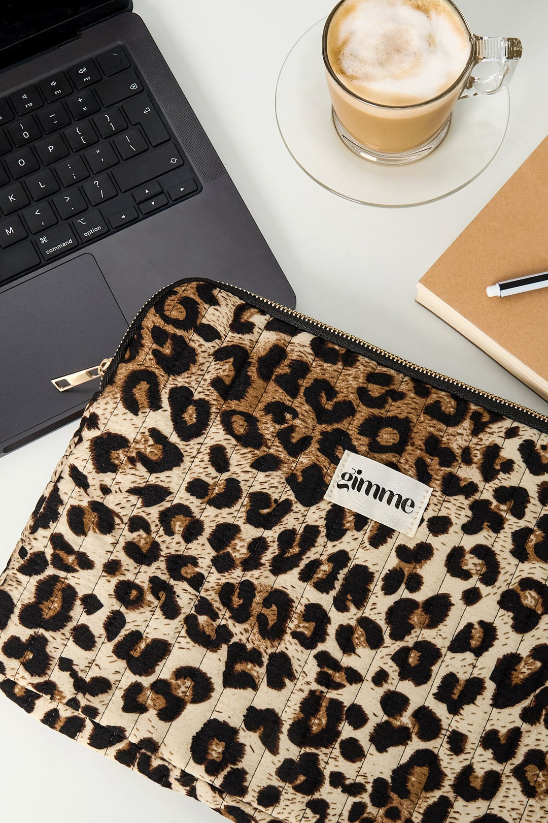 Close-up of leopard print laptop sleeve next to laptop, coffee, and notebook on desk