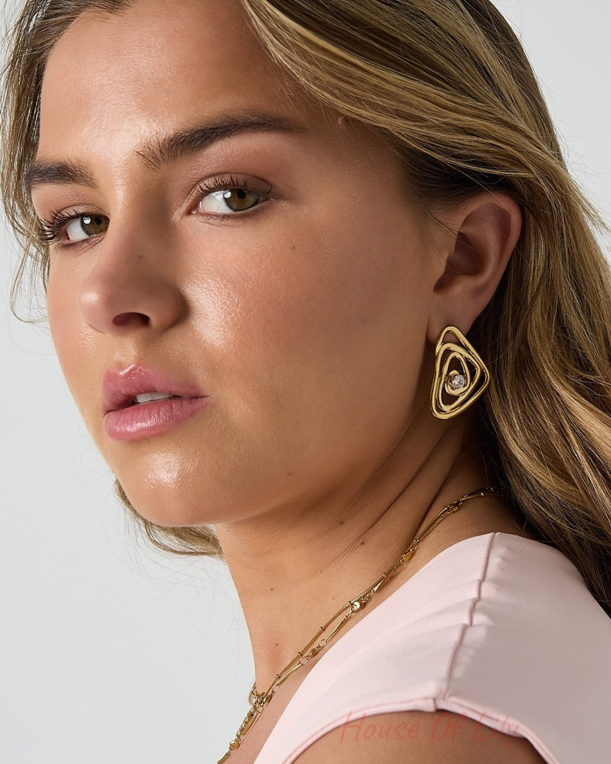 Close-up of a woman wearing gold spiral earrings with rhinestone, styled with delicate gold necklace and light pink top