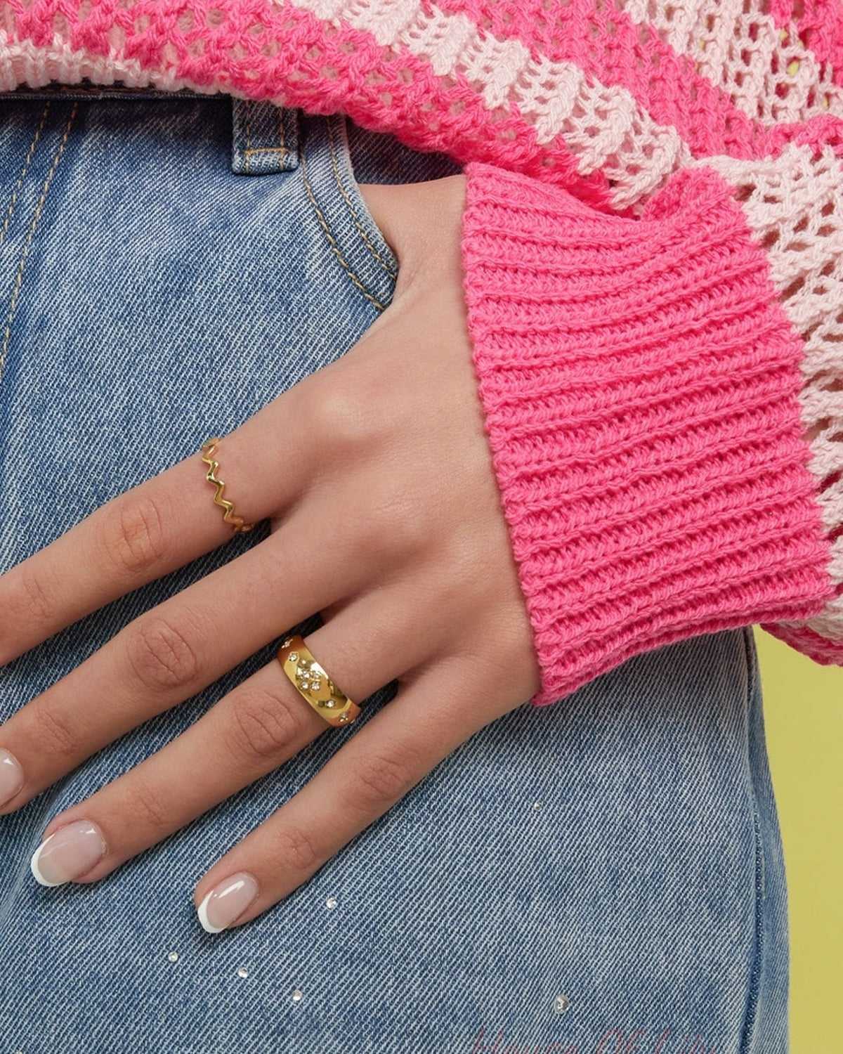 Woman's hand wearing the gold crystal accent ring on the middle finger and a wavy gold ring on the index finger, styled with pink knitwear and denim jeans with rhinestone details