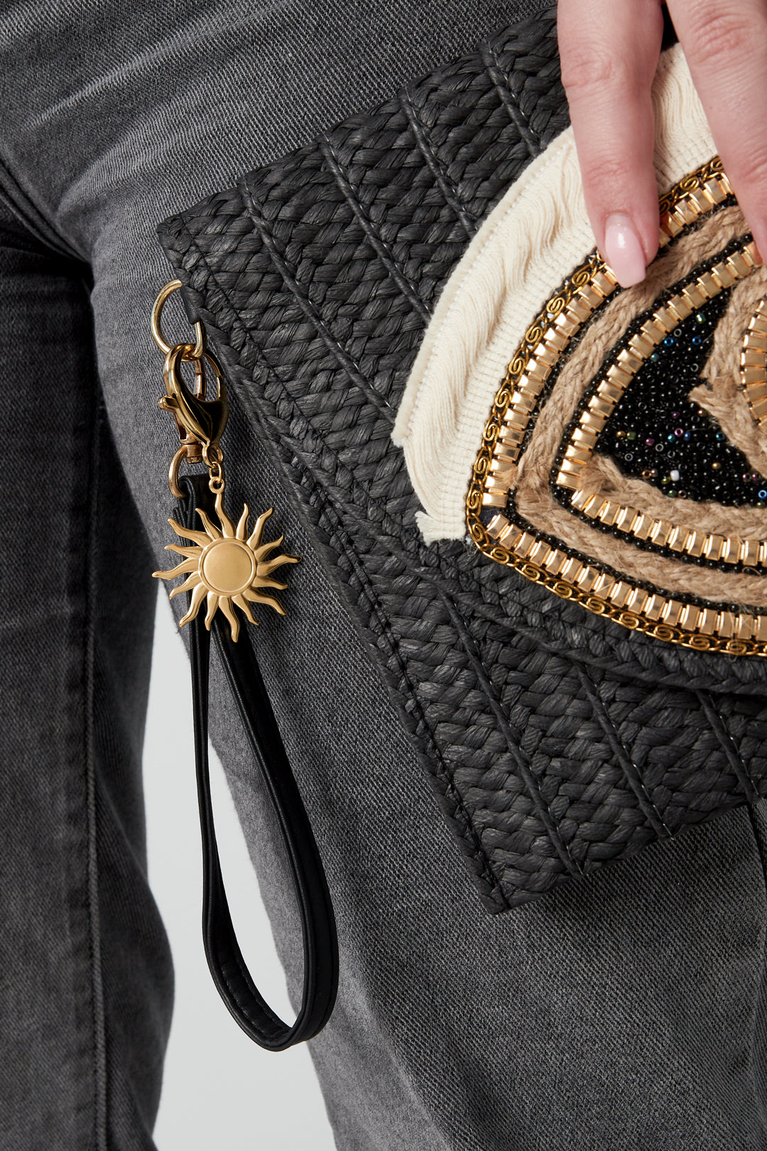 Gold-tone sun keychain charm clipped to a black textured clutch bag held by a person wearing gray pants and showing a manicured hand
