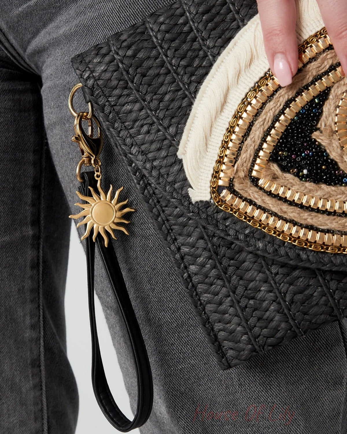 Gold-tone sun keychain charm clipped to a black textured clutch bag held by a person wearing gray pants and showing a manicured hand