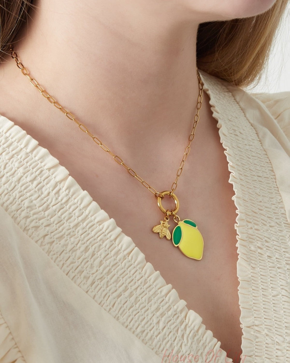 Close-up of a woman wearing a gold chain necklace with a yellow lemon charm and bee detail, paired with a cream blouse