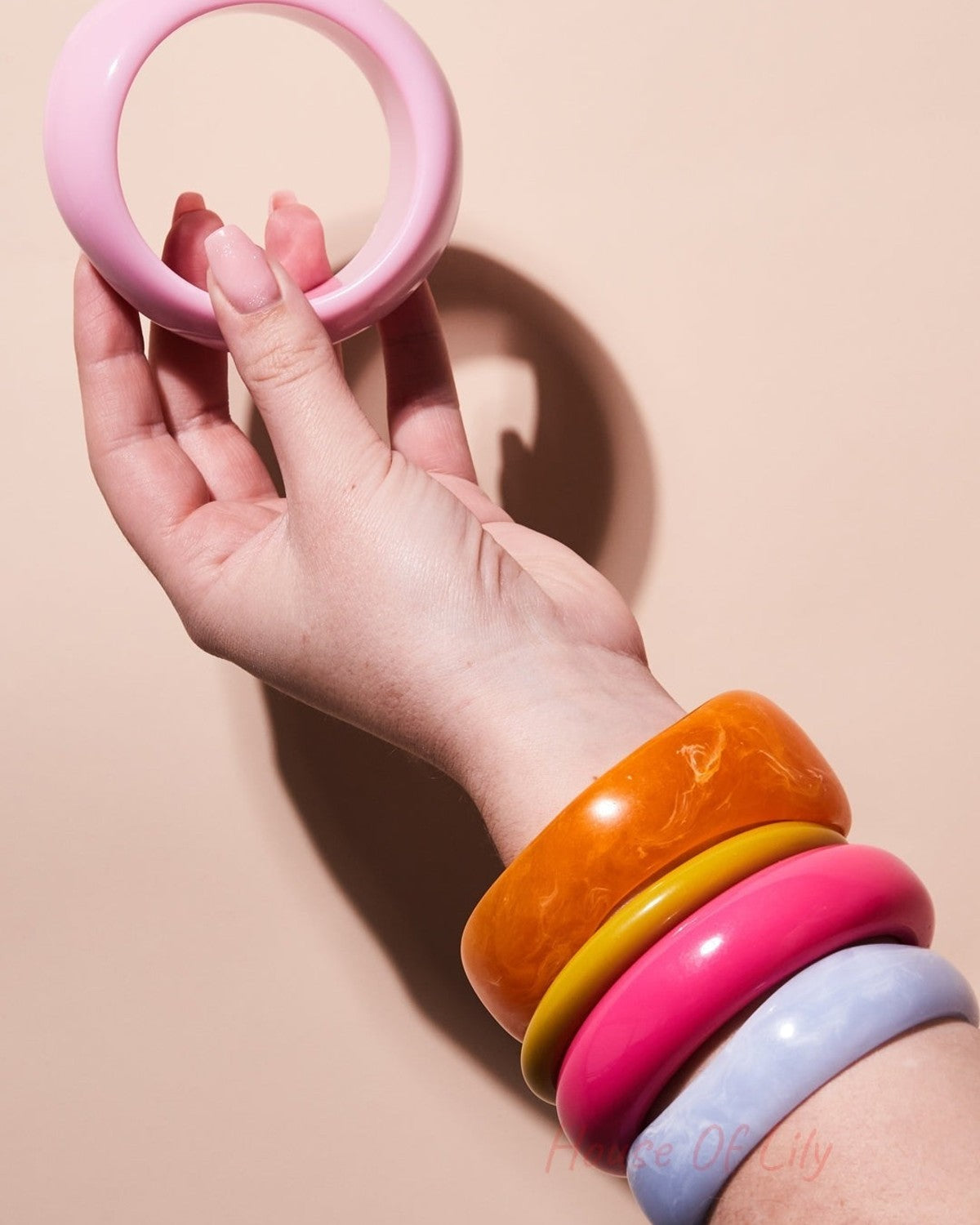 Hand holding a single pink resin bangle against neutral background with arm wearing chunky orange, yellow, pink, and light blue bangles