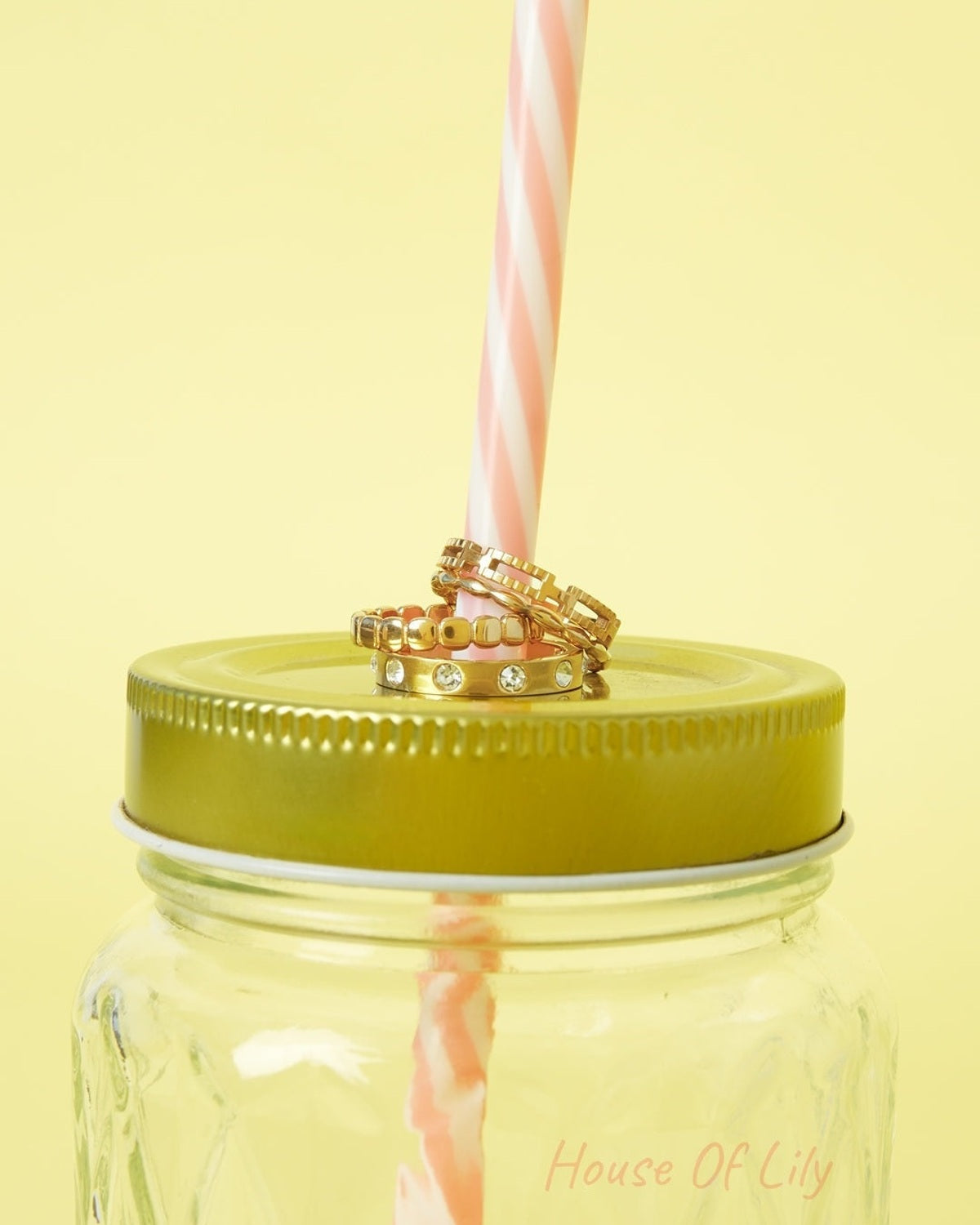 Three gold rings with beaded and gemstone designs stacked on a pink-striped straw in a green lid mason jar on yellow background