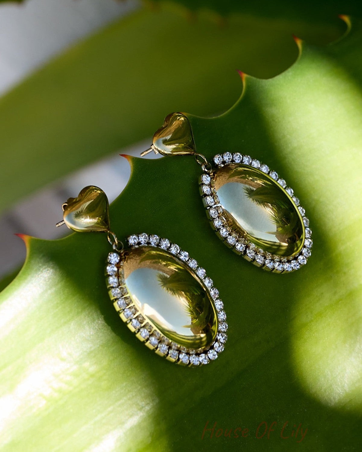 Main Character Drip earrings: gold, oval-shaped drops with crystal halos and heart studs, resting on a green leaf.