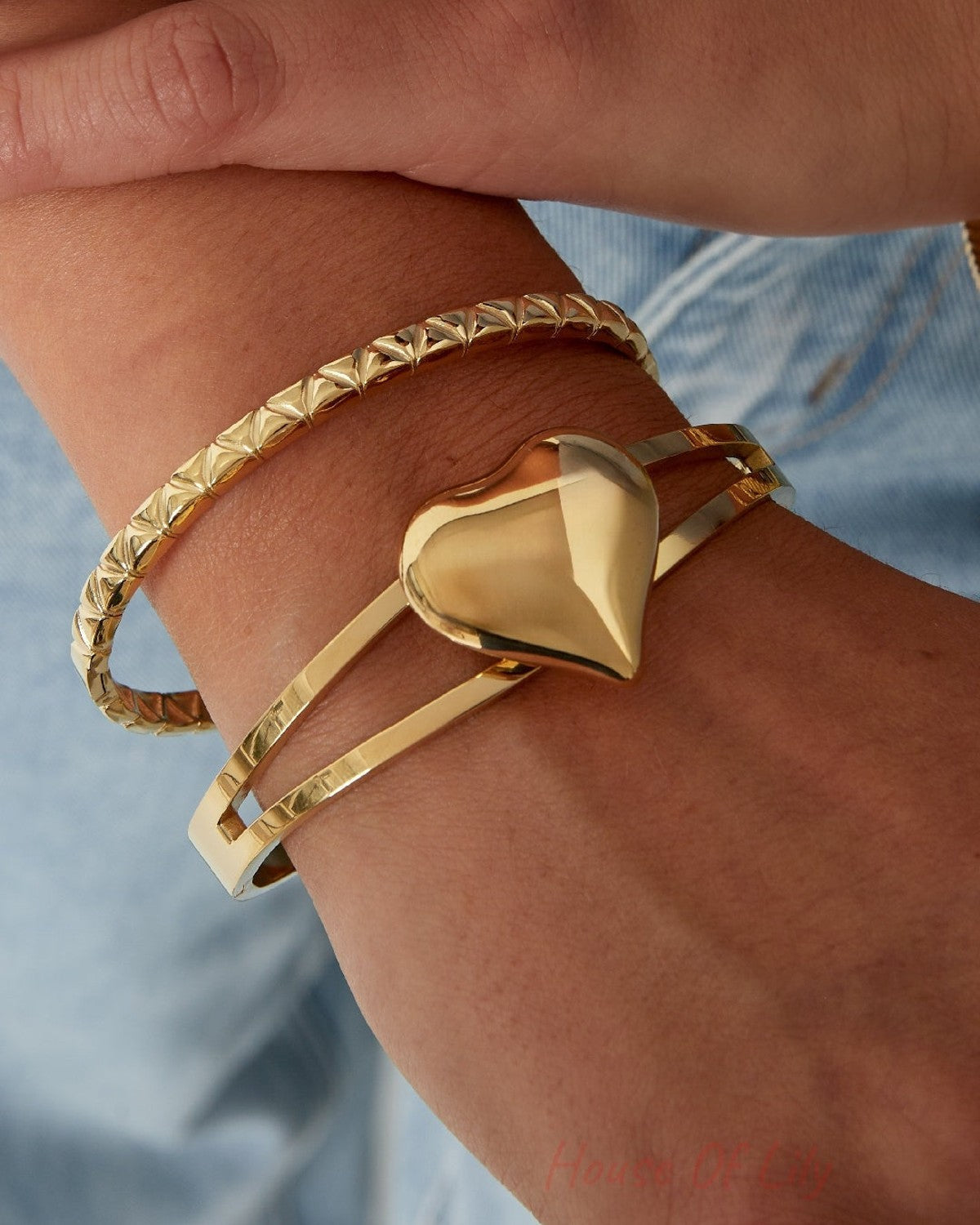 Close-up of wrist wearing a shiny gold double-band heart bangle with a second textured gold bracelet, styled with casual denim