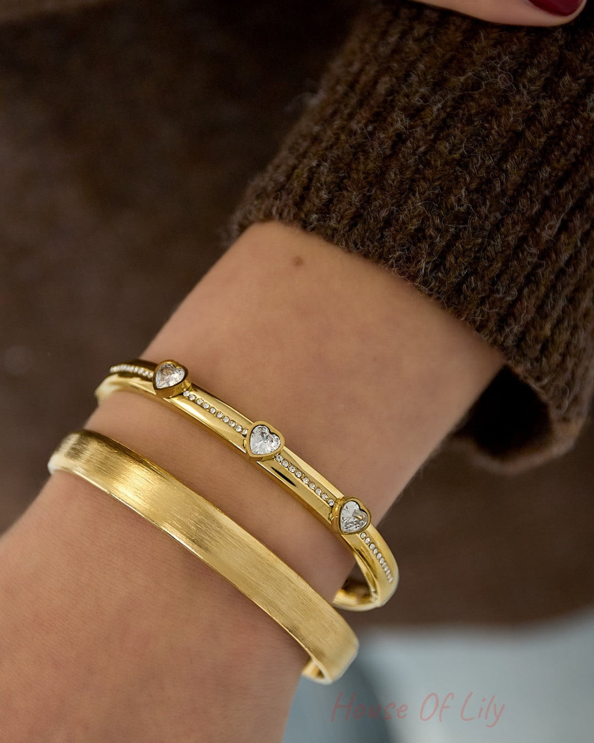 Woman's wrist wearing gold tone heart crystal bangle with a second matte gold bracelet alongside brown knit sweater