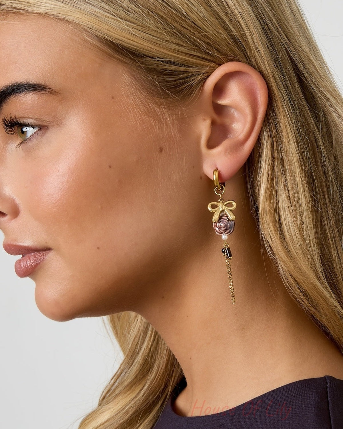 Close-up side view of blonde woman wearing gold-tone floral drop earring with bow, purple flower bead, faux pearls, and chain detail