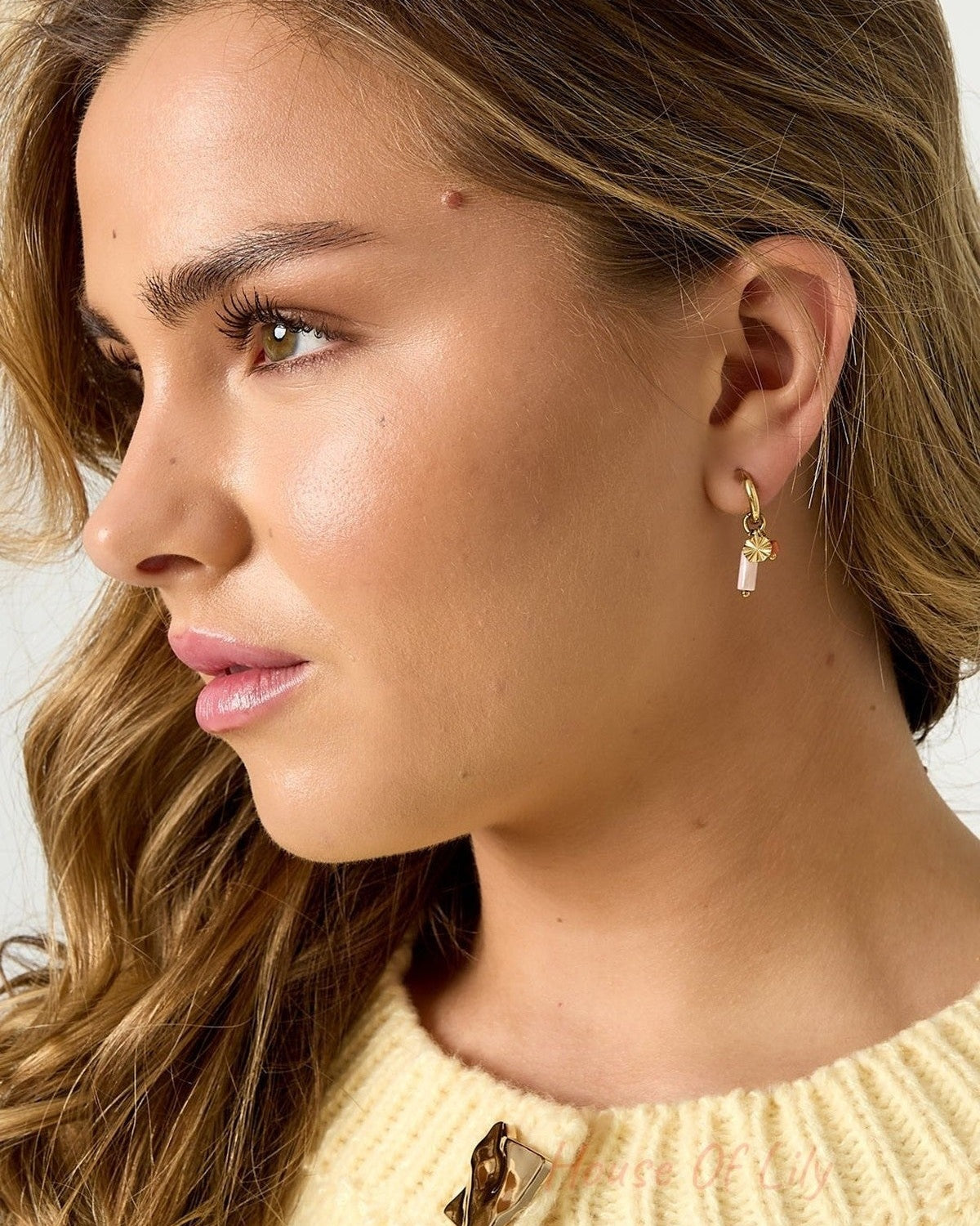Woman with wavy hair wearing gold-tone hooped earrings with pink beads and white stone charms, wearing yellow knitted top