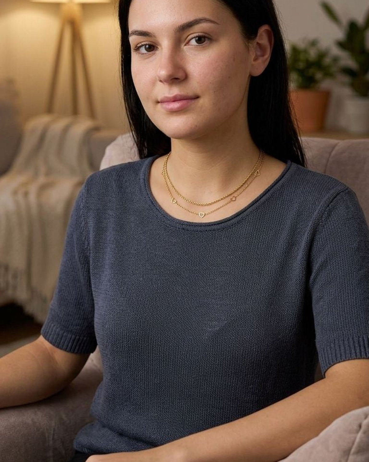 Model wearing a petite gold-plated heart chain necklace on her collarbone in a warm, softly lit indoor setting.