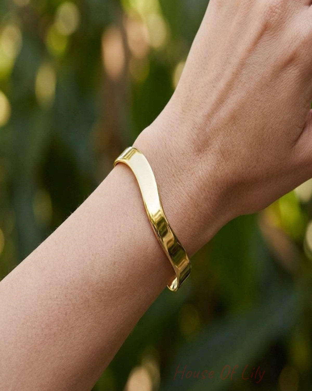 Full-body shot of a female wrist with a small stainless steel cuff bracelet outdoors with greenery, showcasing its delicate design and irregular shape.