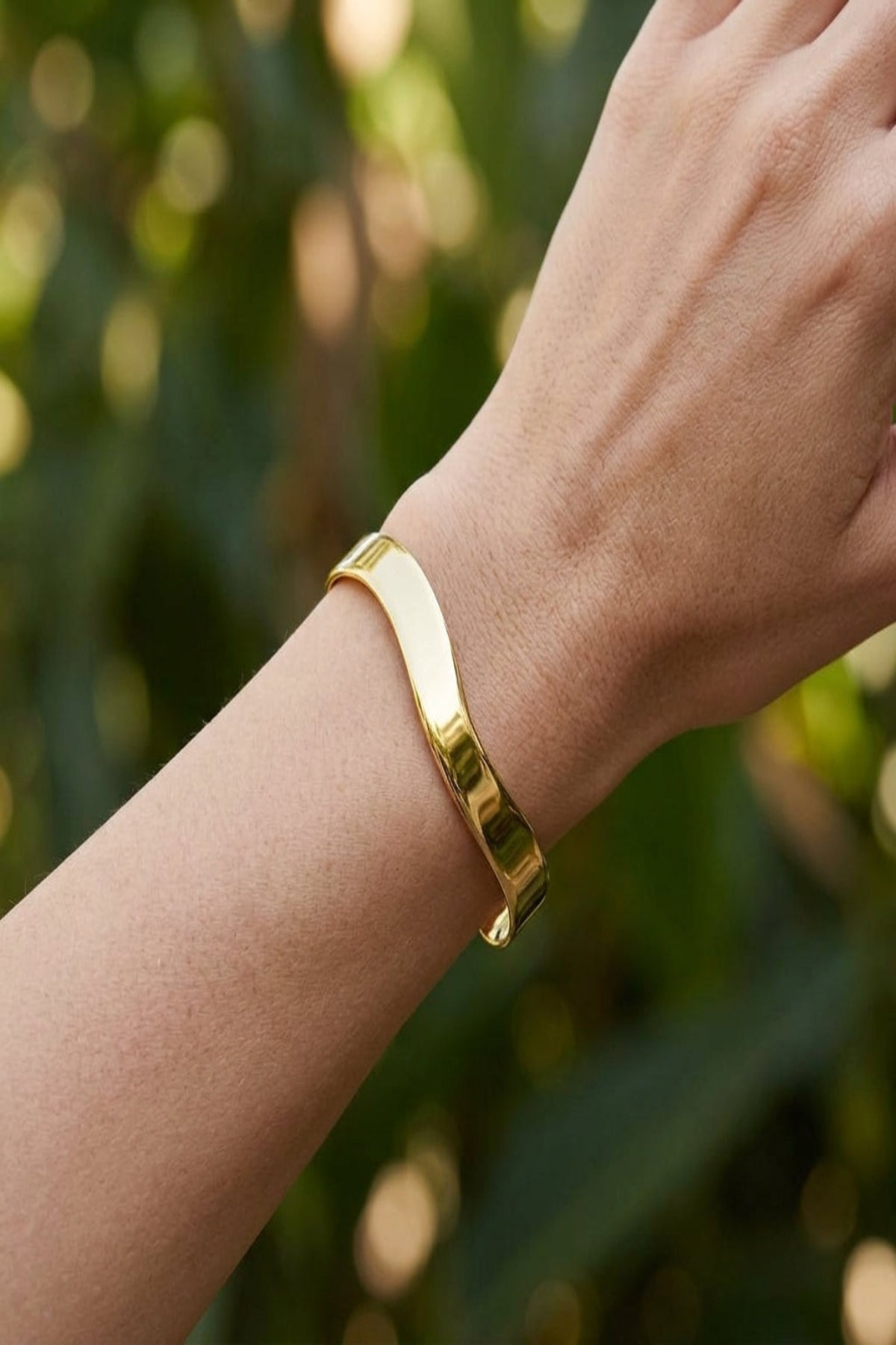 Full-body shot of a female wrist with a small stainless steel cuff bracelet outdoors with greenery, showcasing its delicate design and irregular shape.
