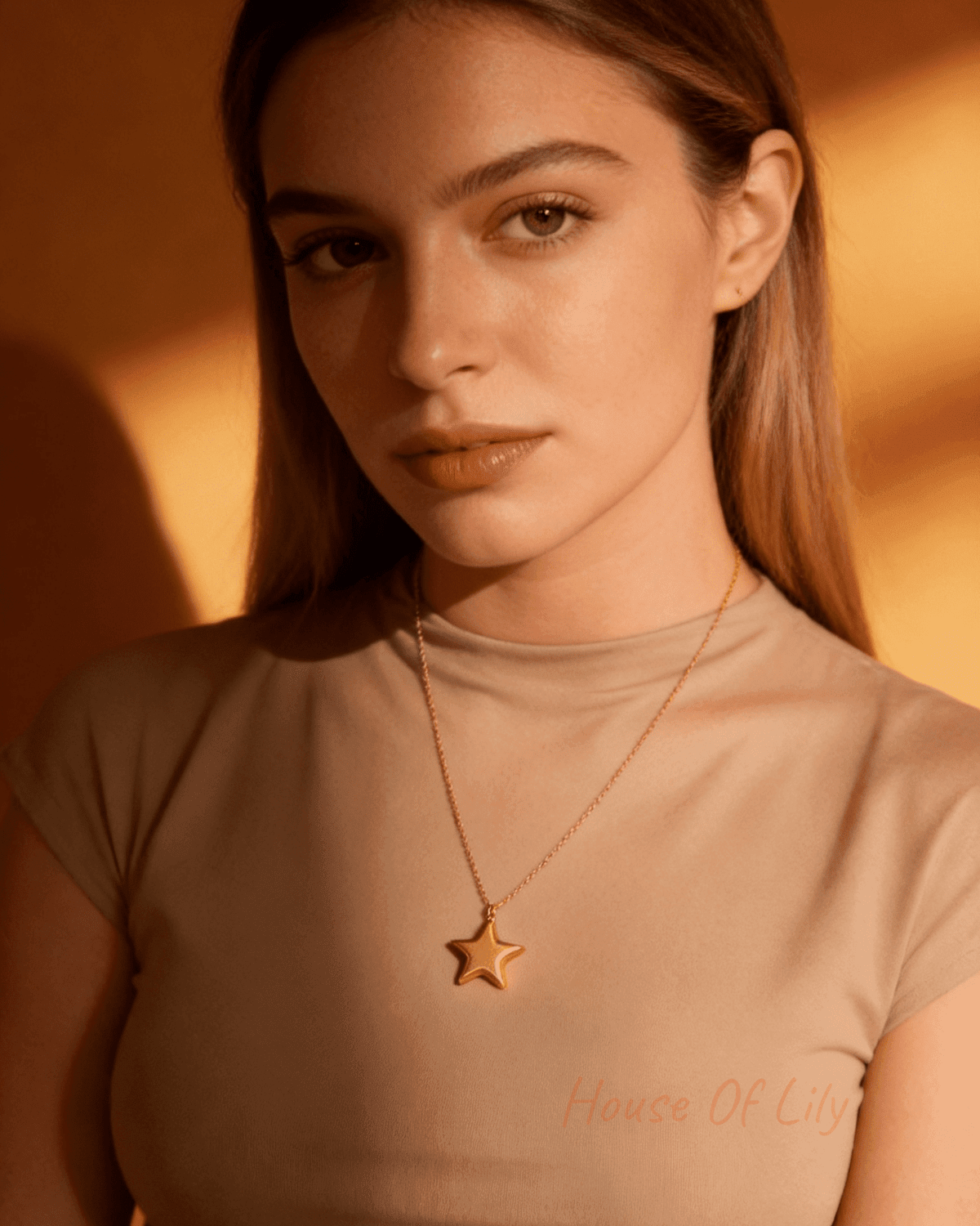 A woman wearing a gold star pendant on a delicate chain against a warm-toned background.