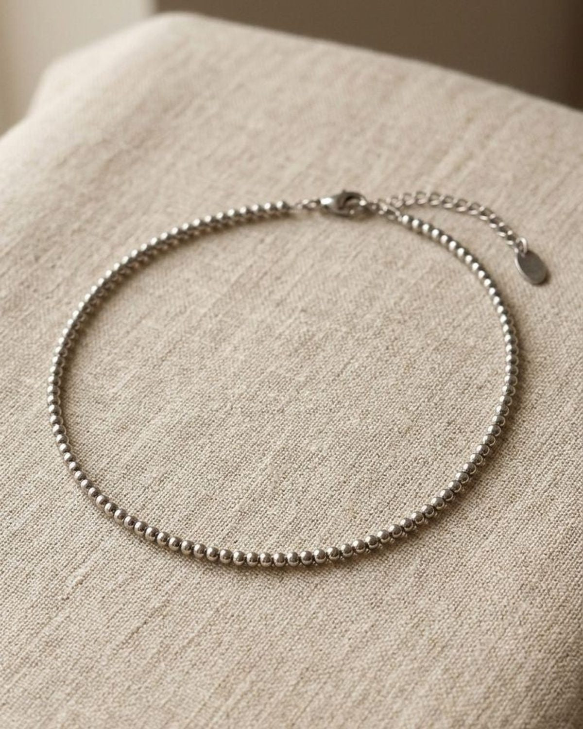 Close-up of delicate stainless steel beaded necklace with 5mm beads on a linen surface, highlighting its subtle shimmer and minimalist design.
