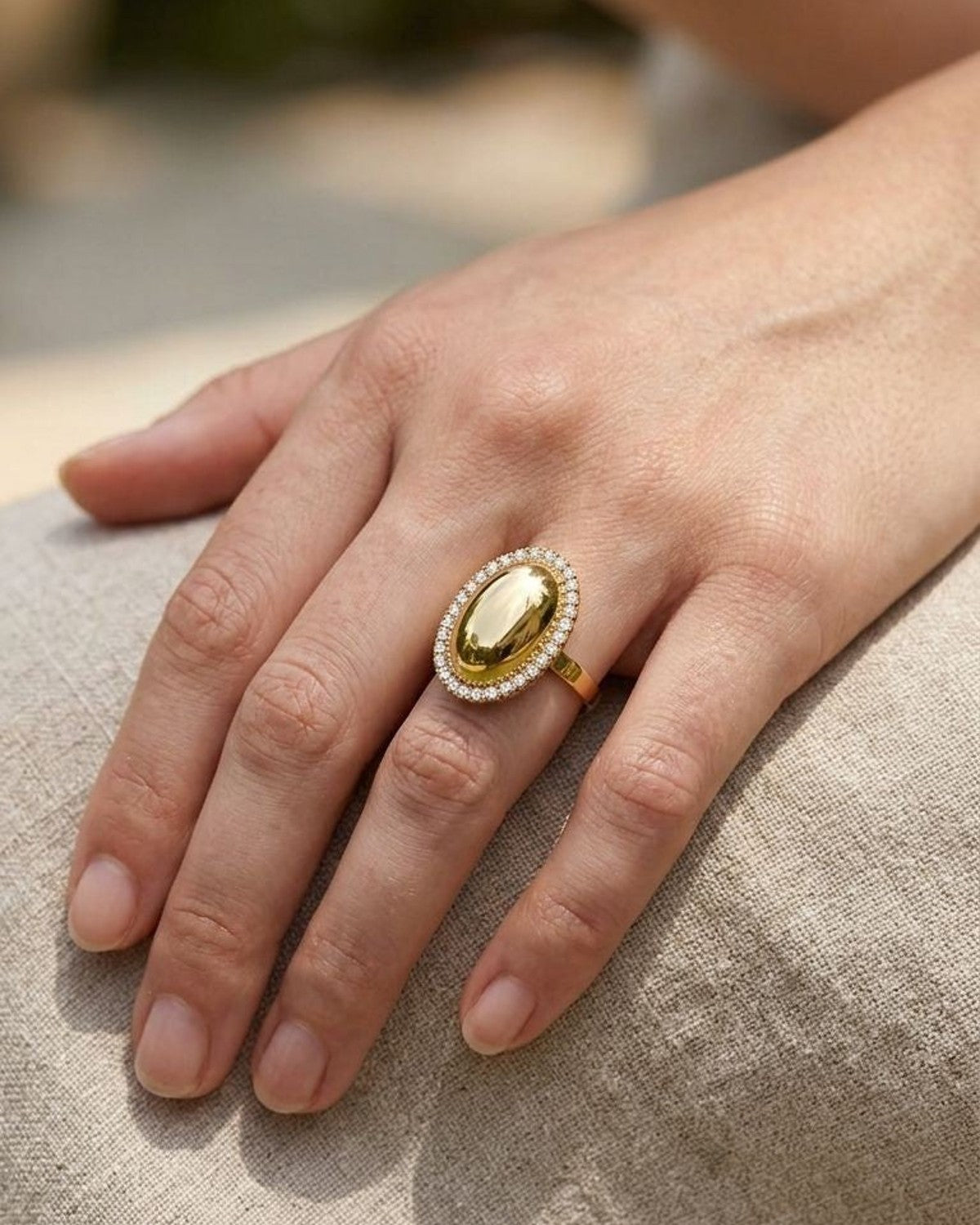 A hand wearing the Golden Oval Statement ring on the middle finger, showing its sparkle and delicate design against a rustic fabric background.