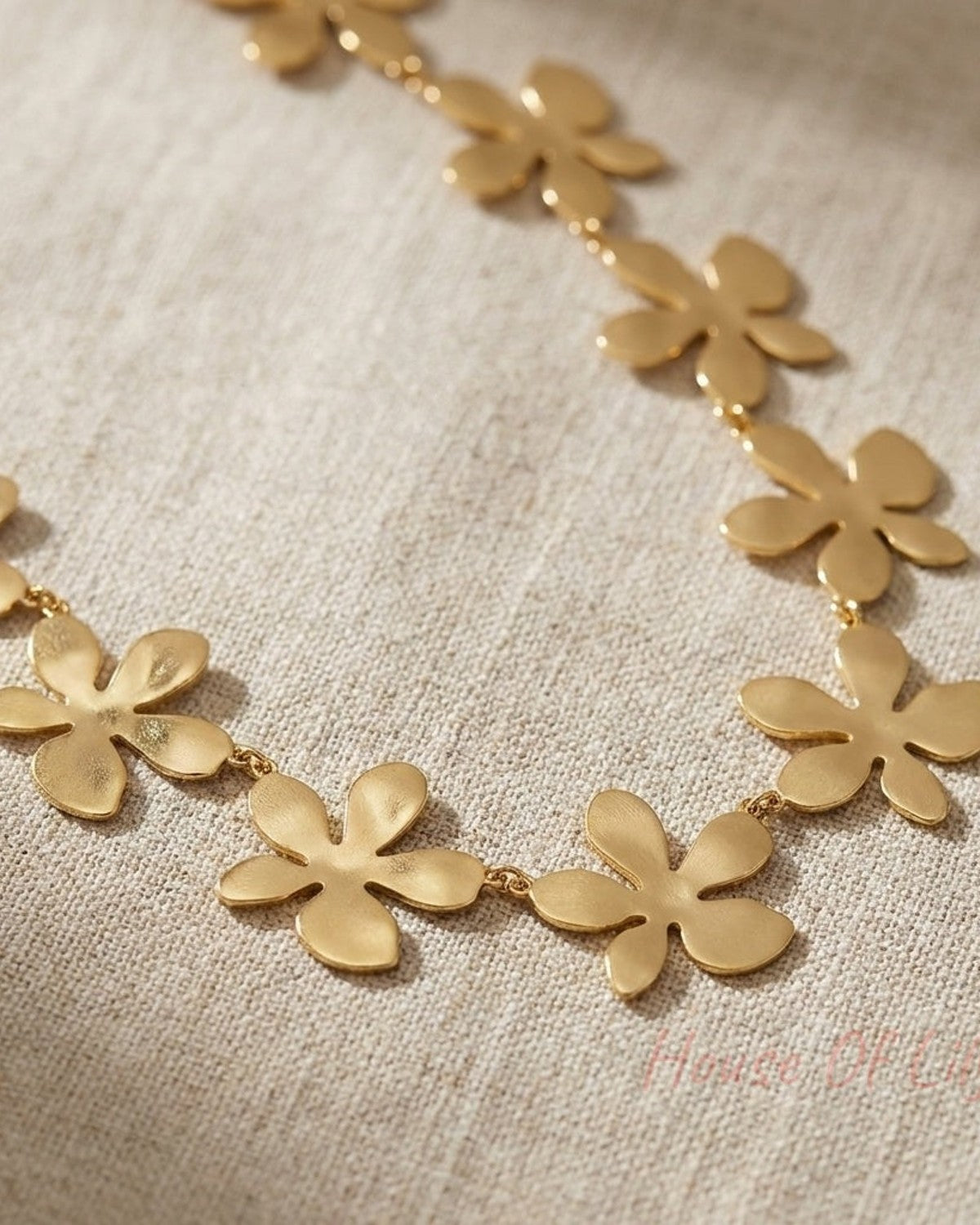Detailed close-up of a gold flower necklace with micro-texture, draped on beige fabric, showcasing delicate craftsmanship and subtle shine.