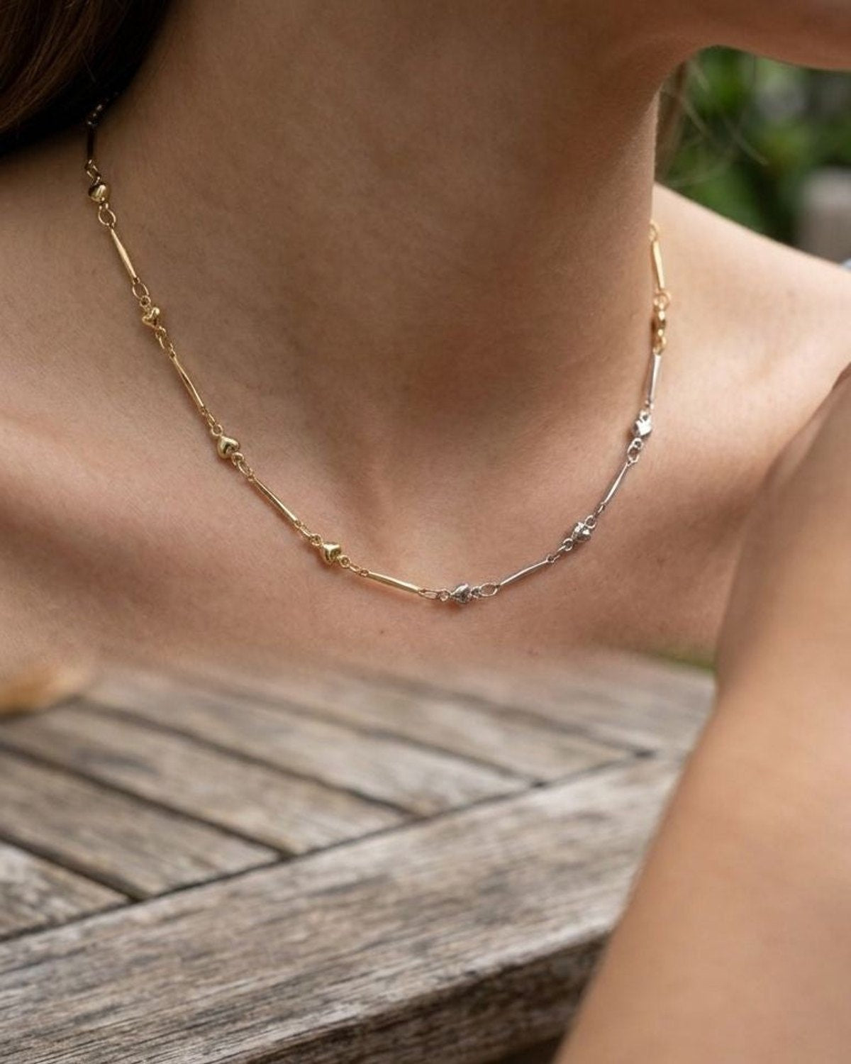 Close-up of the polished love link necklace on a model's neck, outdoors on a rustic wooden table with greenery blur, emphasizing its delicate links.