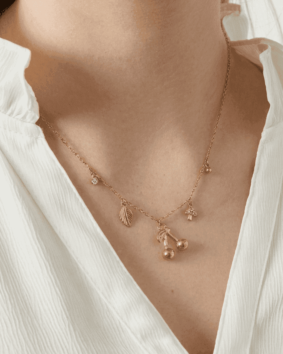 Gold chain necklace featuring delicate charms of cherries, a leaf, a mushroom, and a small crystal, worn on a neck.