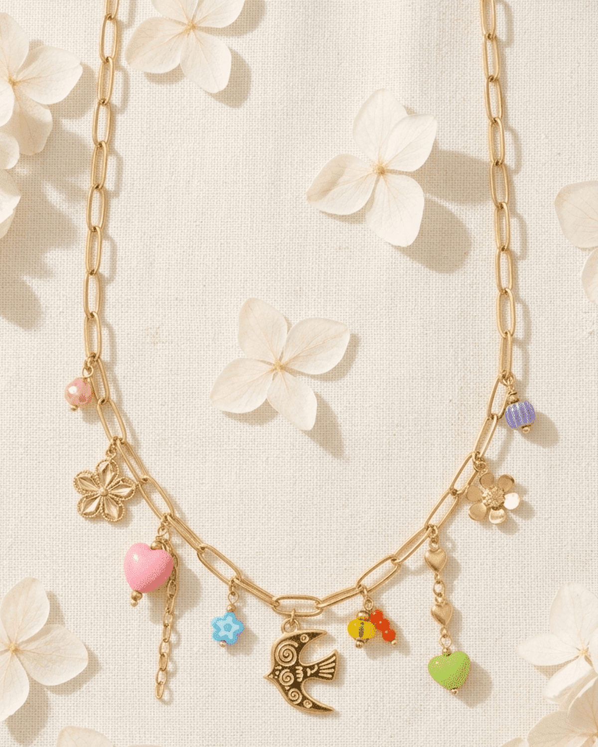Gold link chain necklace with assorted colorful charms, including a bird, hearts, and flowers, on a white floral fabric.