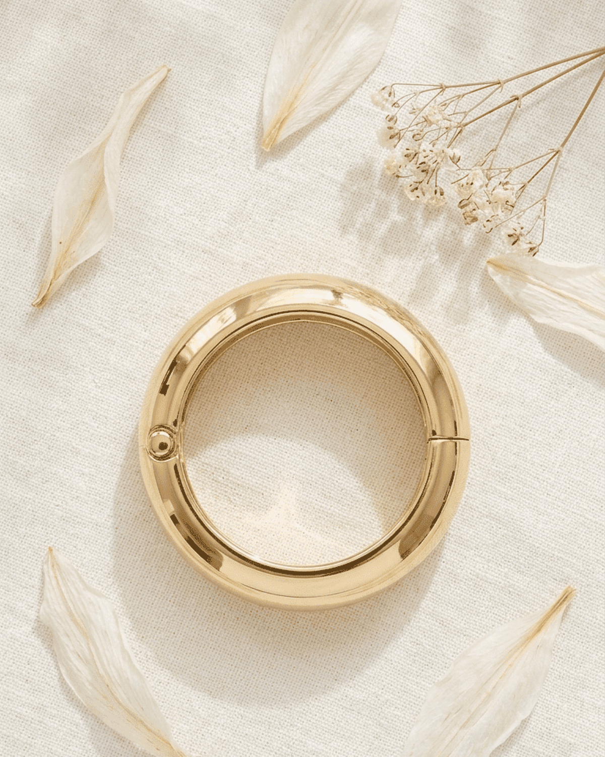 Chunky gold hinged bangle on a textured white background with scattered flower petals and baby's breath.