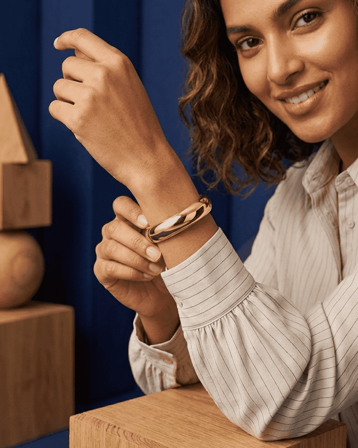 Chunky gold bangle with a polished, smooth finish worn on the wrist of a smiling woman in a striped shirt.