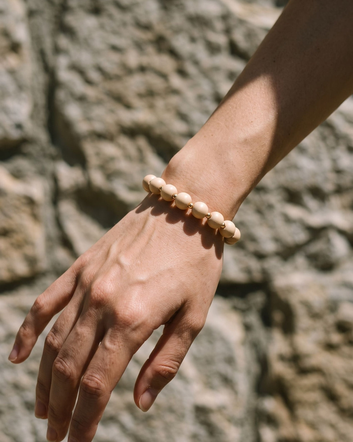 A slender person wearing a petite brown and pink wooden beaded bracelet against a textured stone wall background.