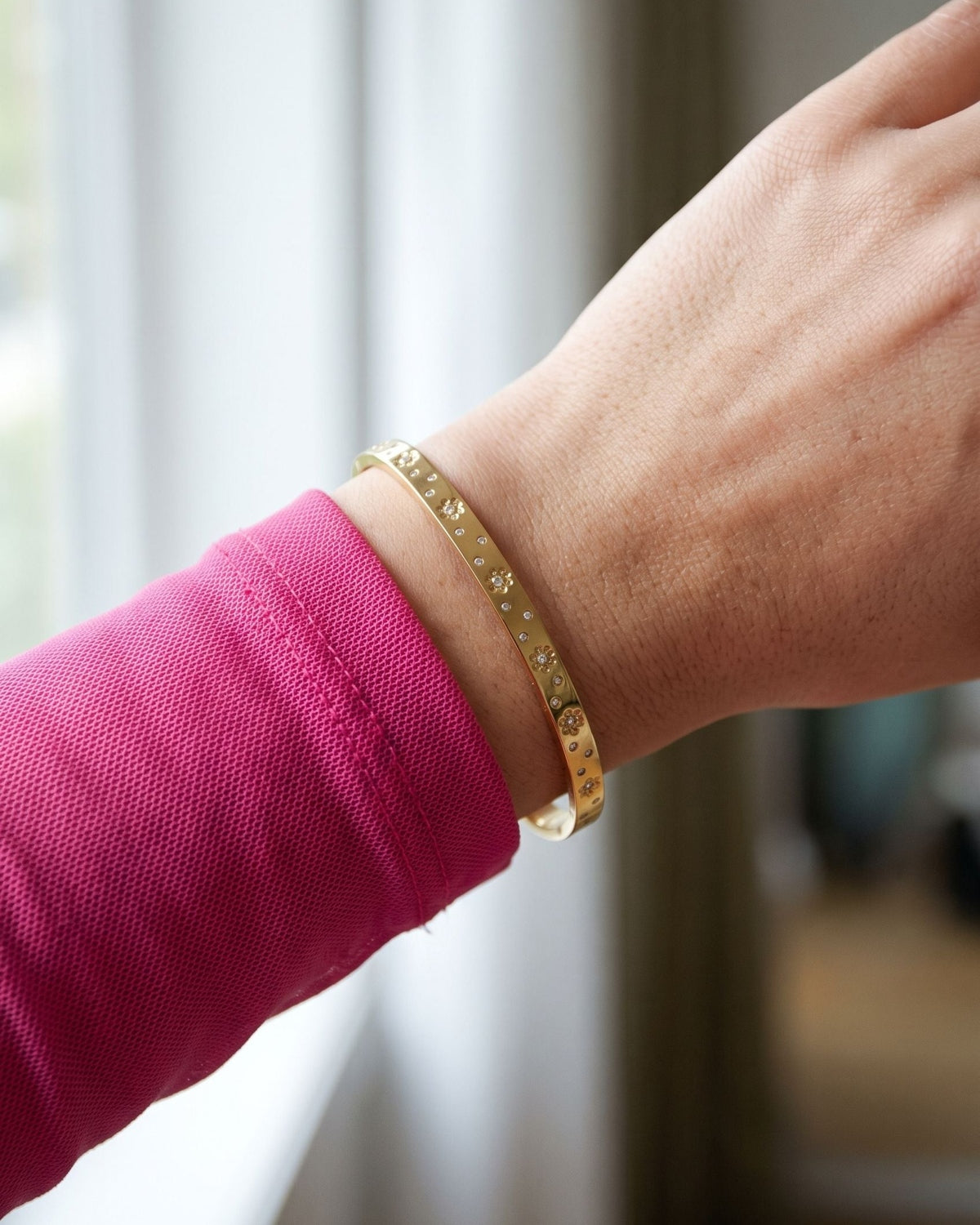 A slim, petite gold-toned flower bangle worn on a wrist over a bright fuchsia long-sleeved mesh top.