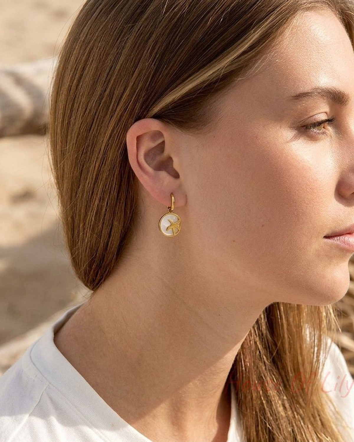Close-up of starfish glow hoops earrings with shell details worn on a model's ears, highlighted by natural sunlight with beach tones in background.