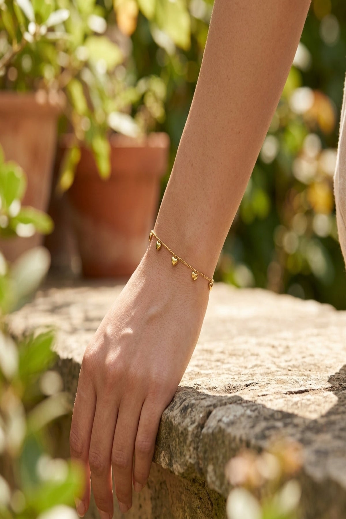 Model wearing a subtle gold-colored stainless steel bracelet with heart charms outdoors, natural daylight.