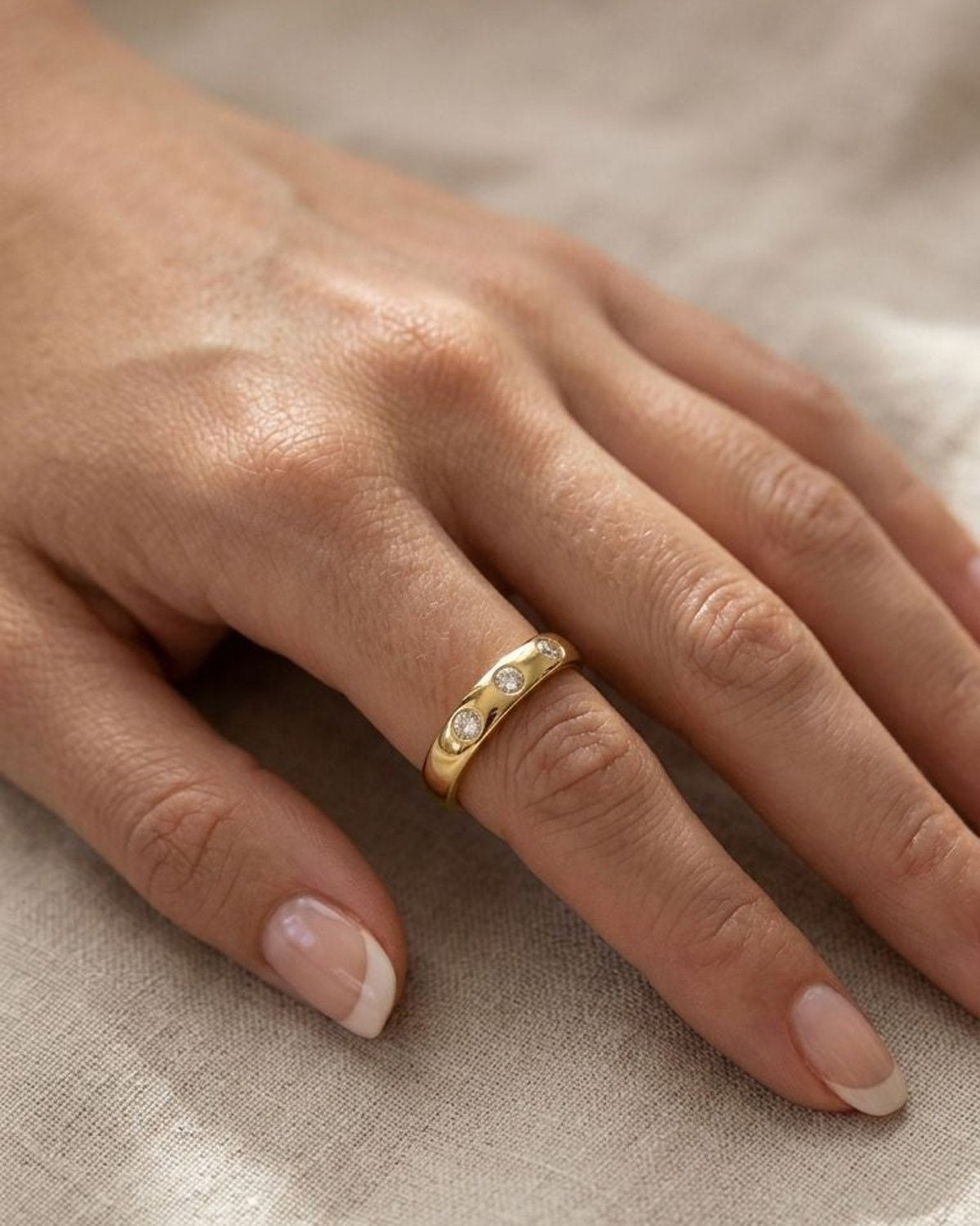 Woman wearing the petite gold ring with three zircon stones on her finger, resting on a neutral linen cloth, showcasing its elegant design.